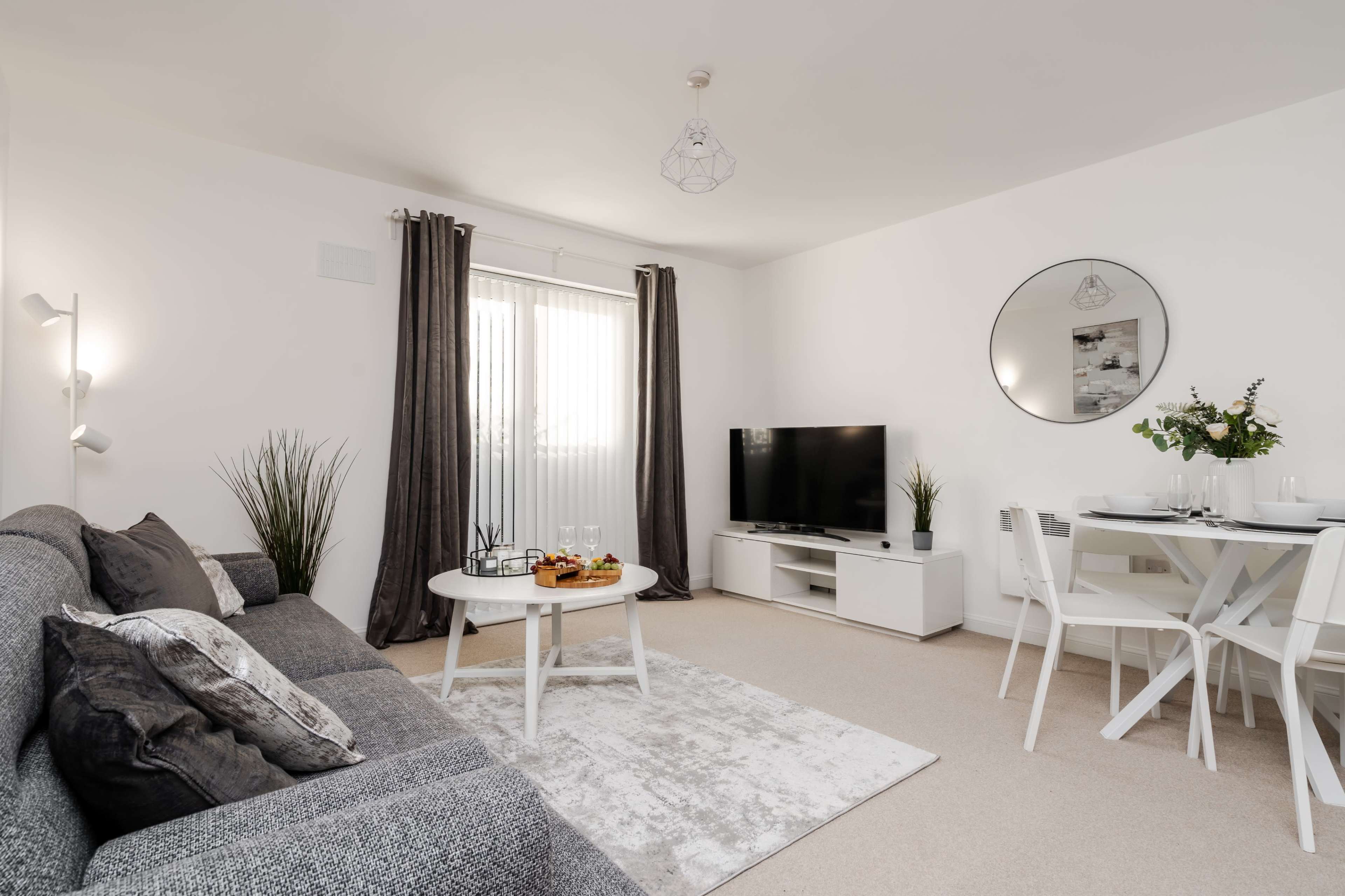A modern living room features a gray sofa, a round coffee table with snacks, a flat-screen TV on a white unit, and a dining table set for four, all illuminated by natural light through sheer curtains.