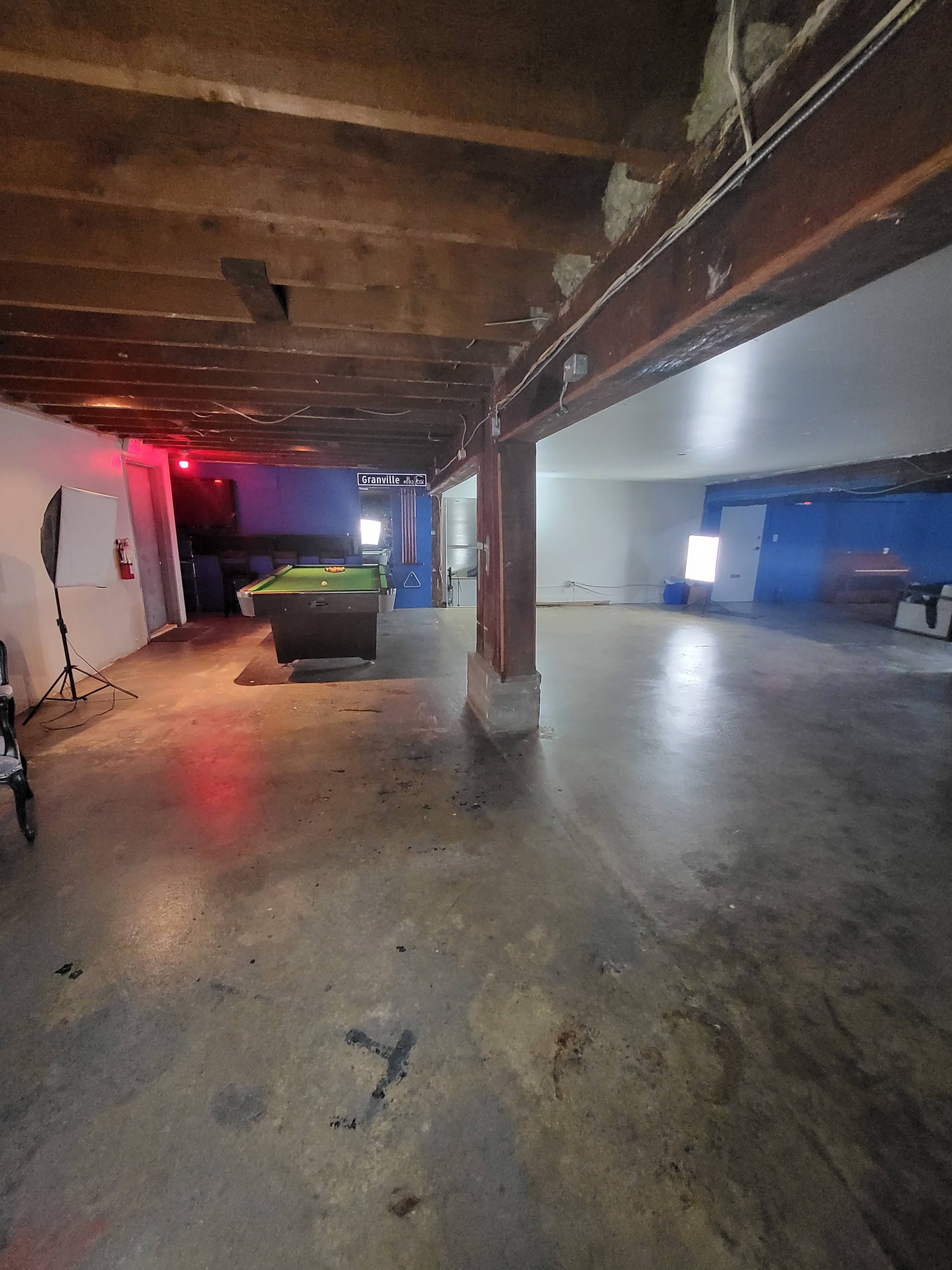 A spacious basement area with exposed wooden beams, a pool table in the center, and concrete flooring.