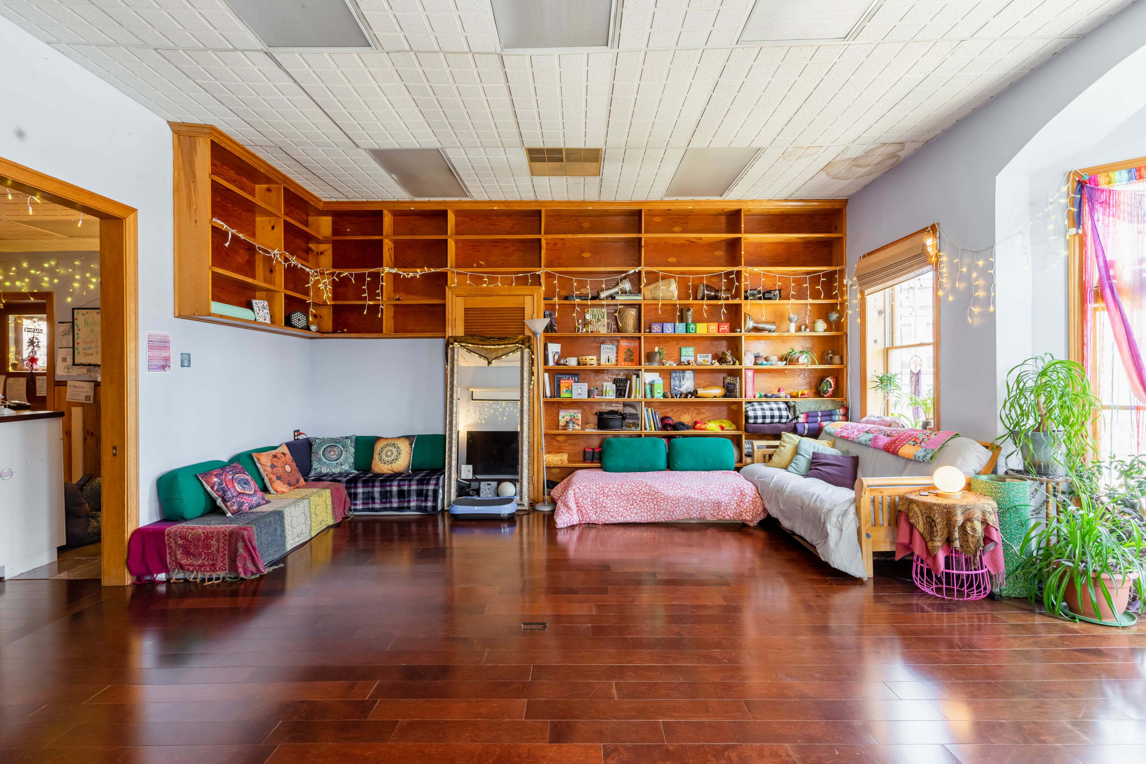 A spacious interior with a wooden floor, a wall of bookshelves filled with various items, and several couches and pillows arranged for seating.