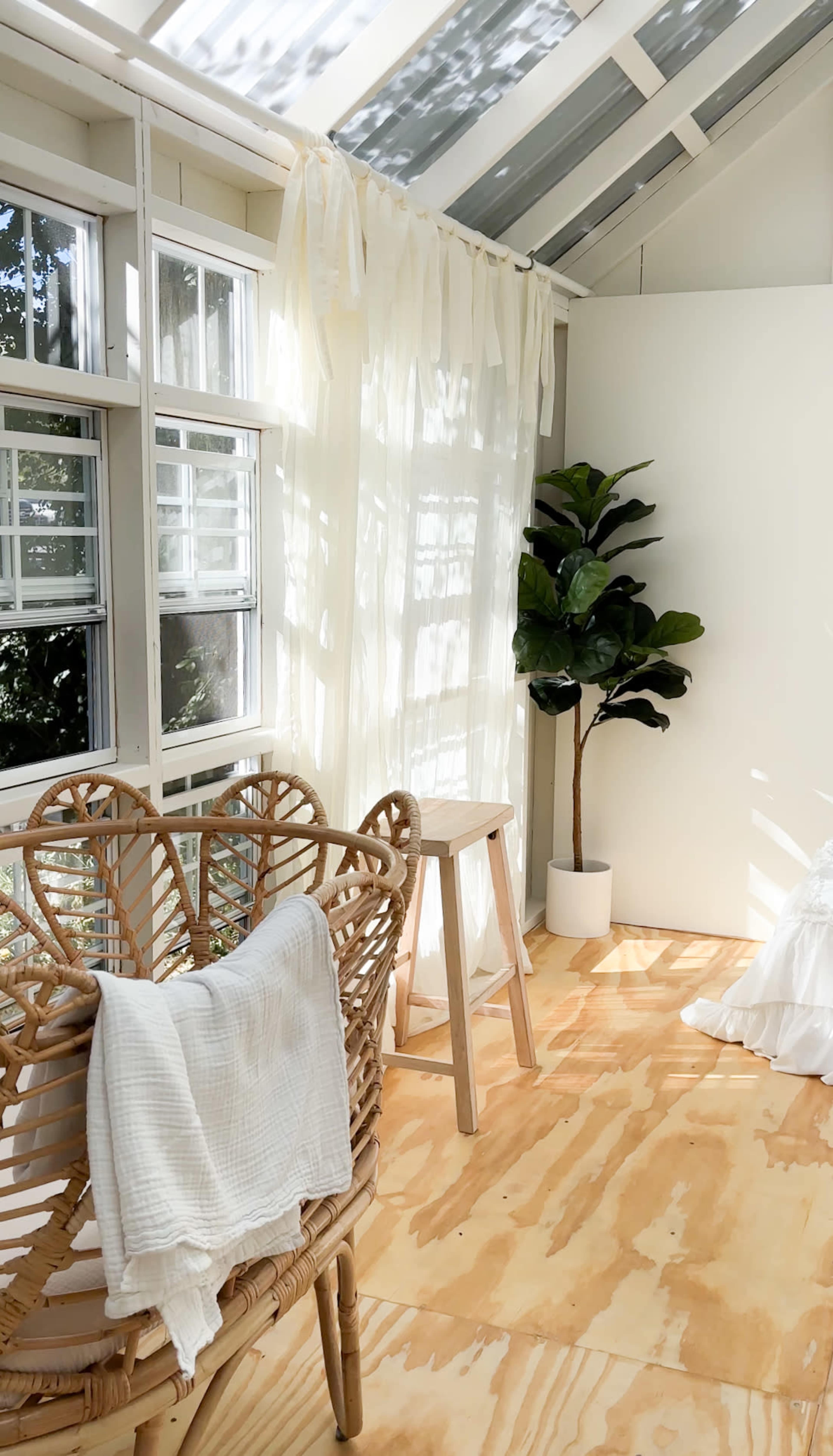 The image shows a bright, sunlit room featuring a rattan chair, a small wooden stool, and a potted plant, with sheer curtains allowing natural light to filter in.