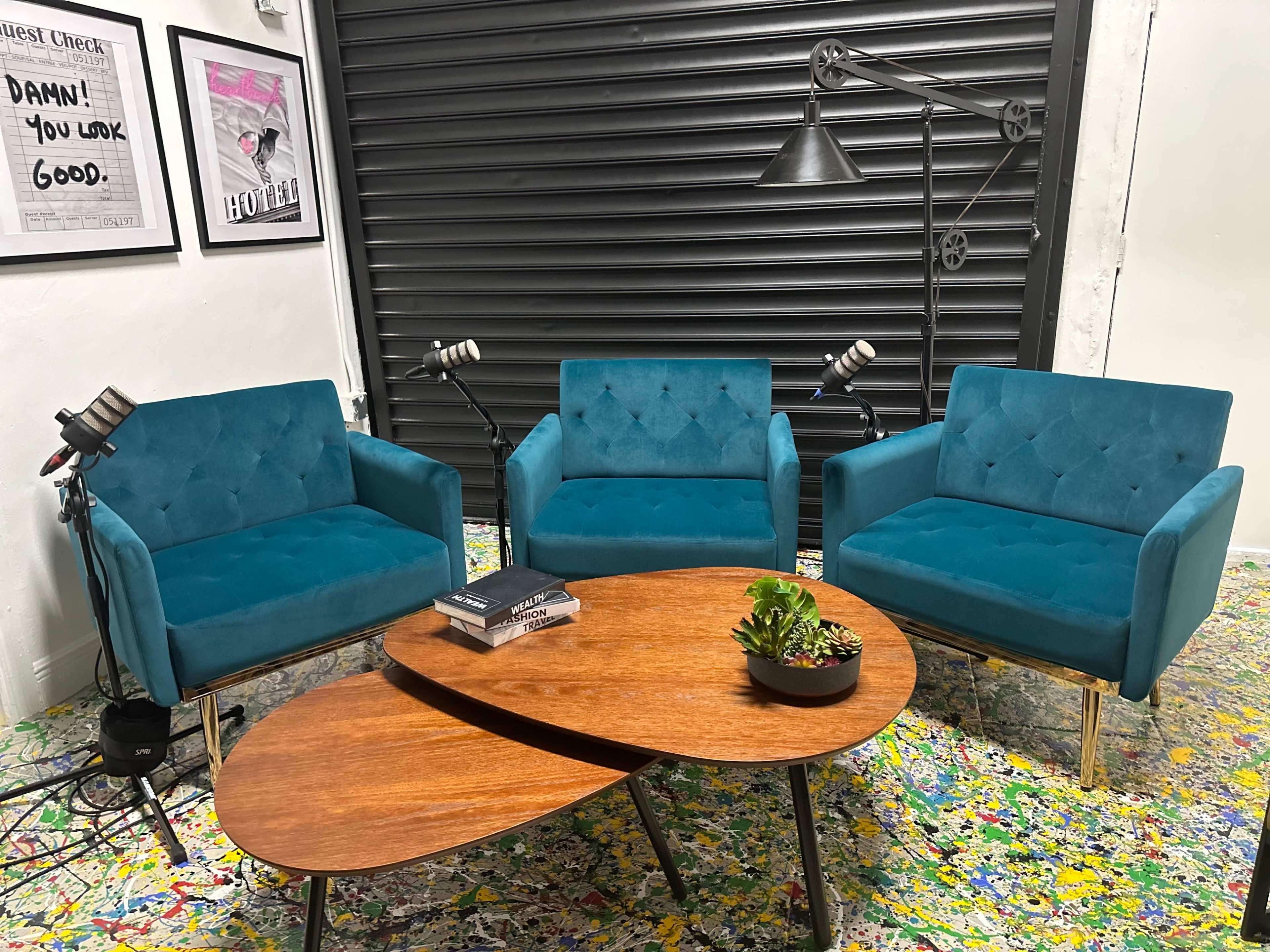 Three teal upholstered chairs arranged around a wooden coffee table on a colorful splattered floor, with a wall-mounted lamp and framed artwork in the background.