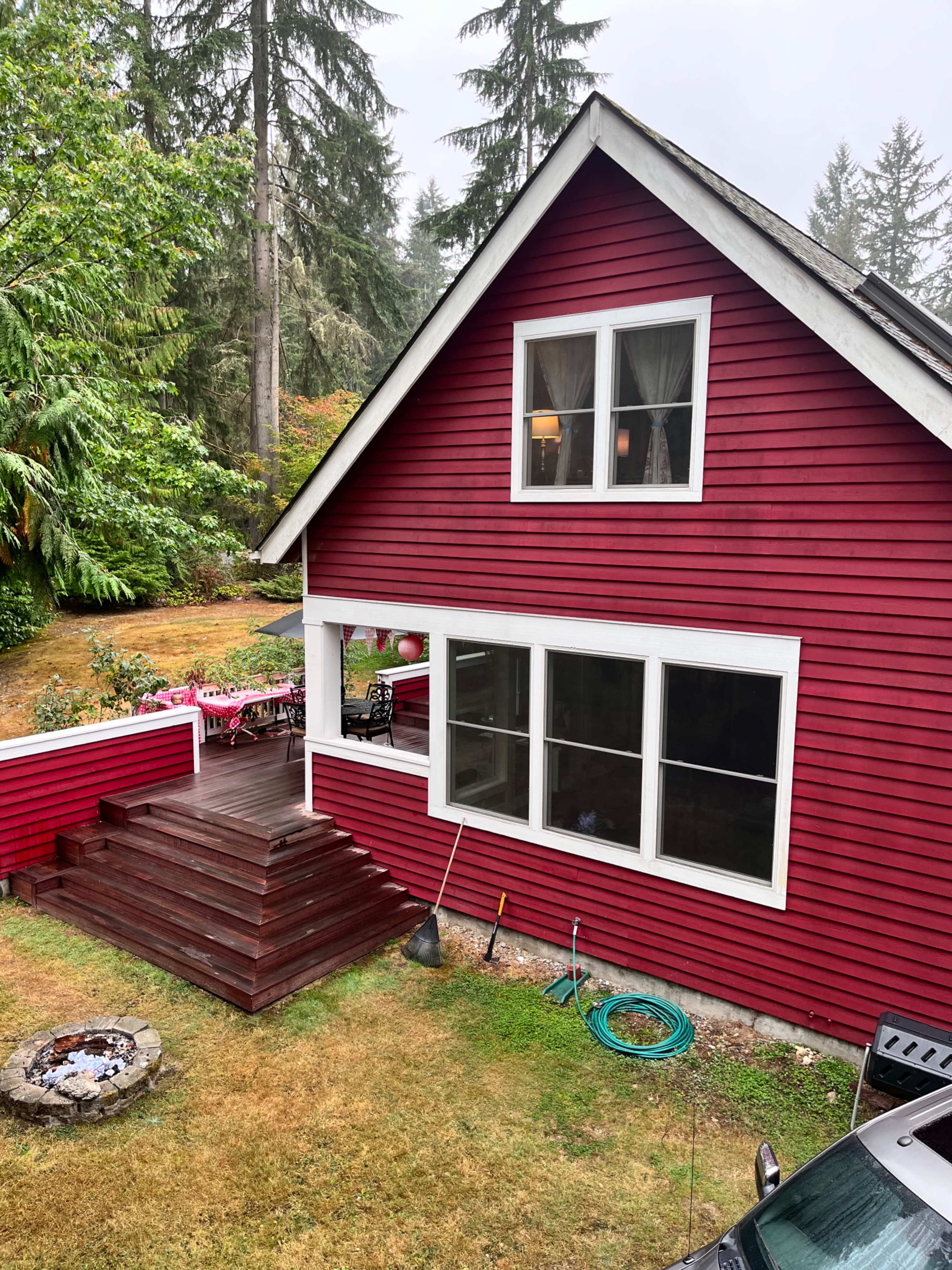 A red house with white trim features a wooden deck and large windows, surrounded by trees and a grassy yard with a fire pit.