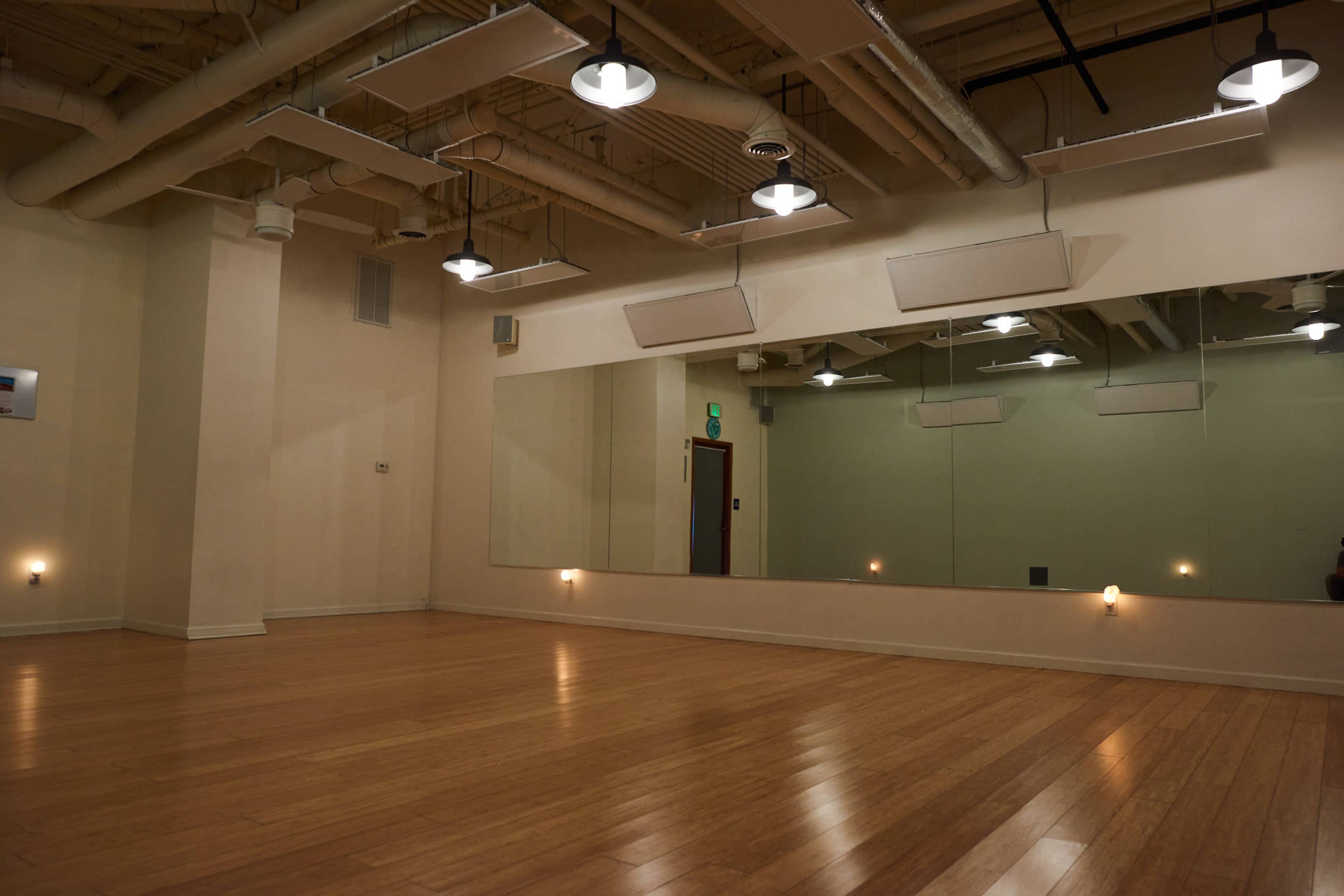 A large, empty studio with wooden floors and a wall of mirrors under overhead lights.