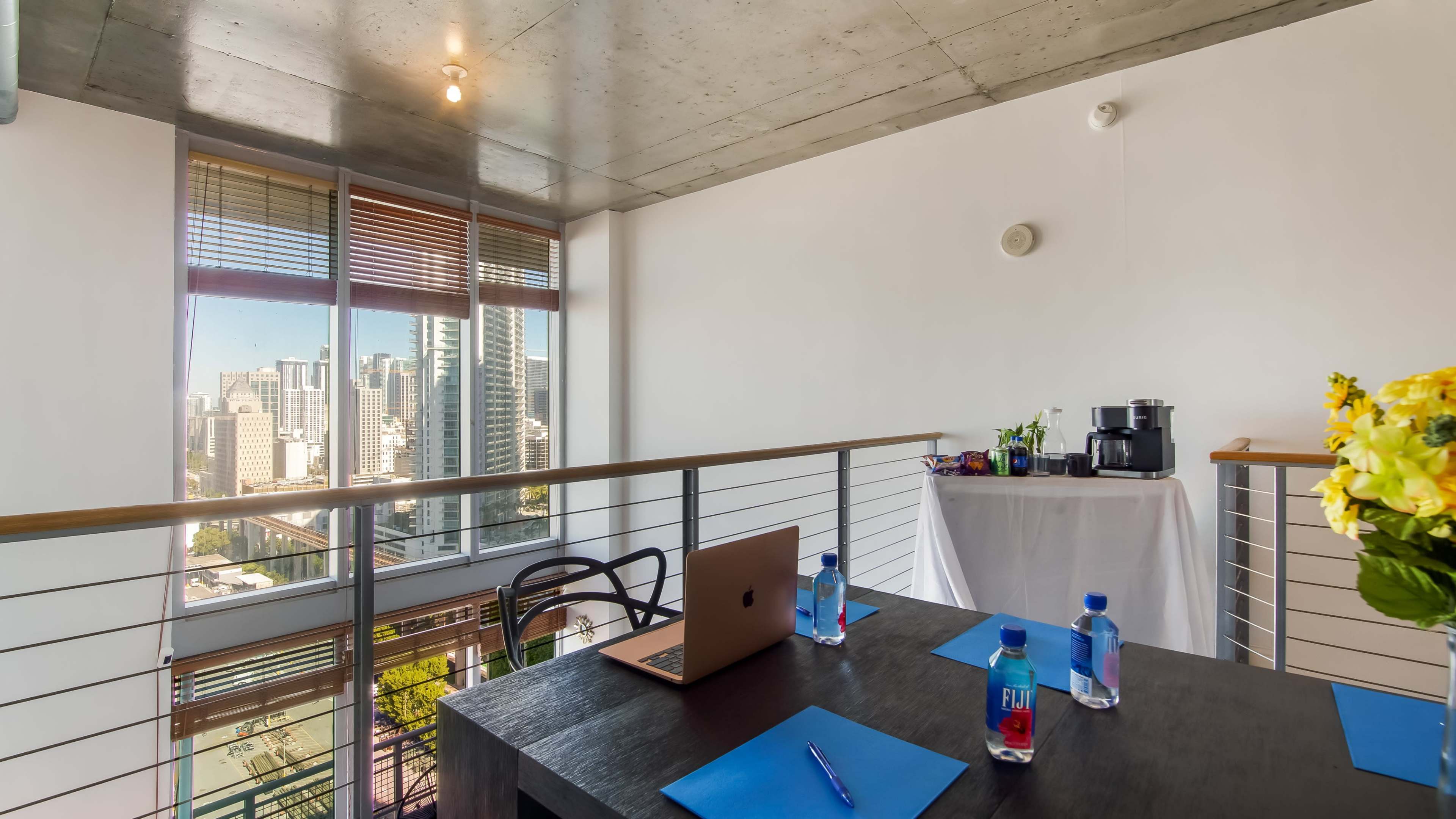 A modern workspace features a table with a laptop and water bottles, set against a backdrop of large windows overlooking a city skyline.