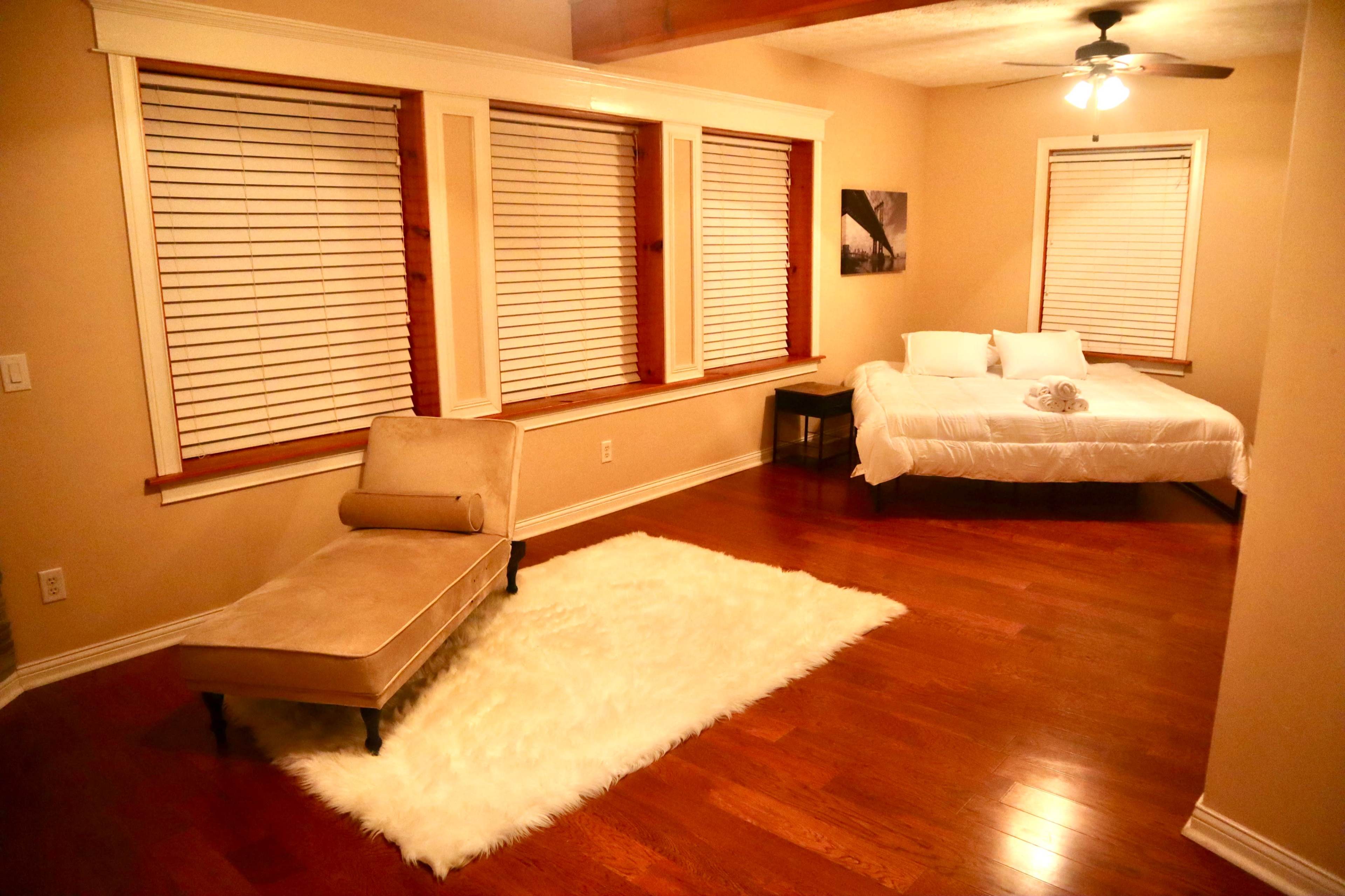 The room features a bed, a chaise lounge, and a large area rug on hardwood flooring, with windows covered by blinds.