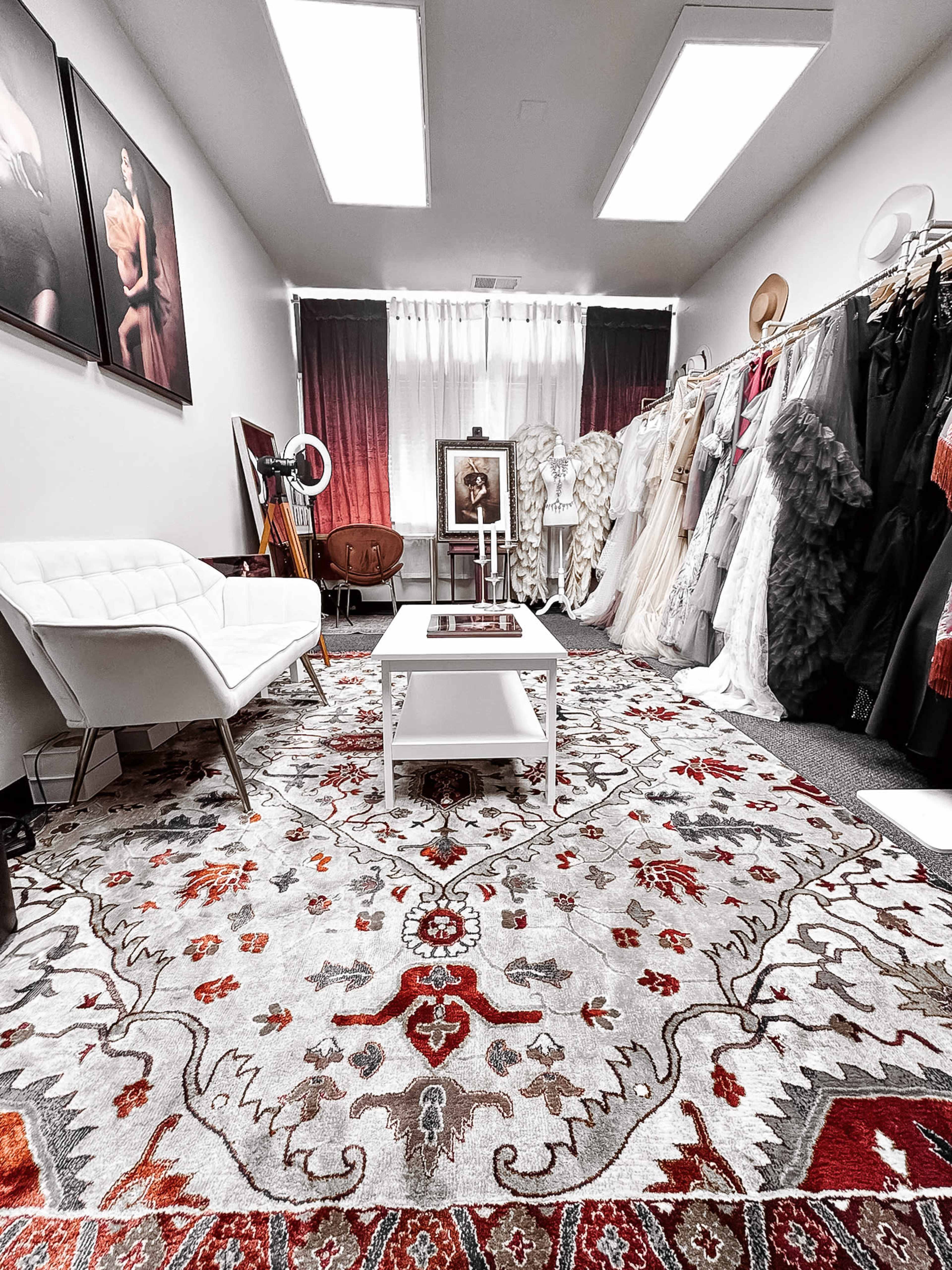 The image shows a well-lit boutique interior featuring a seating area with a white chair and a coffee table, surrounded by clothing racks displaying various dresses and a patterned rug.