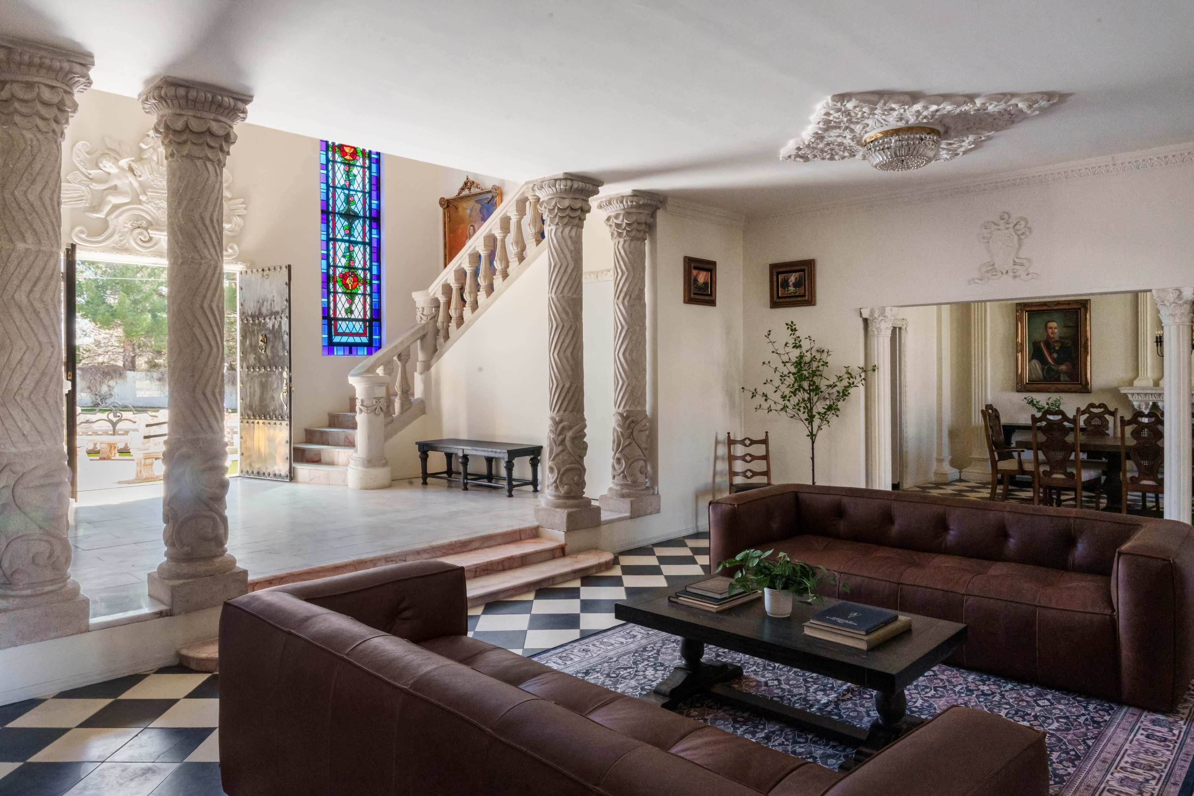 The image depicts a spacious interior living area featuring two brown leather sofas, decorative columns, a staircase leading to an upper level, and a large stained glass window.