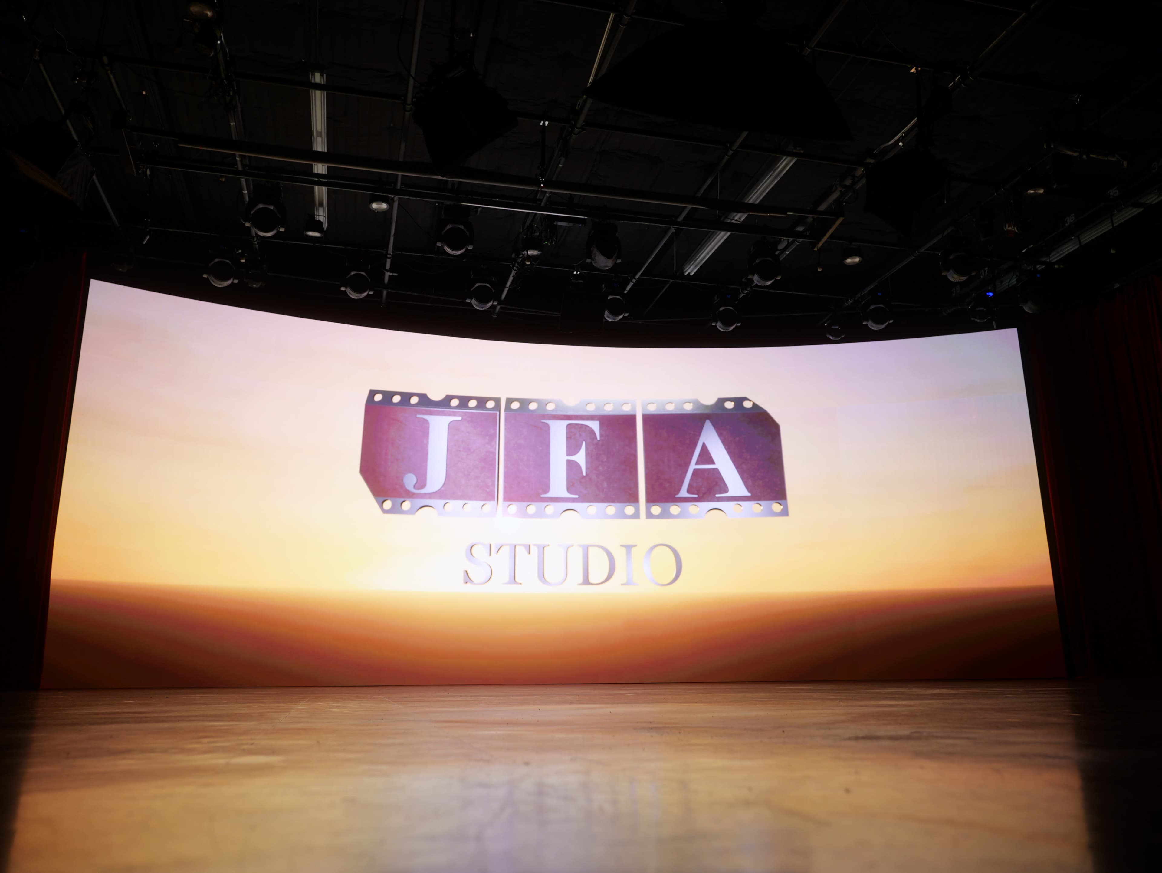 A large, illuminated screen in a studio setting displays the text "JFA STUDIO" over an orange sunset background.