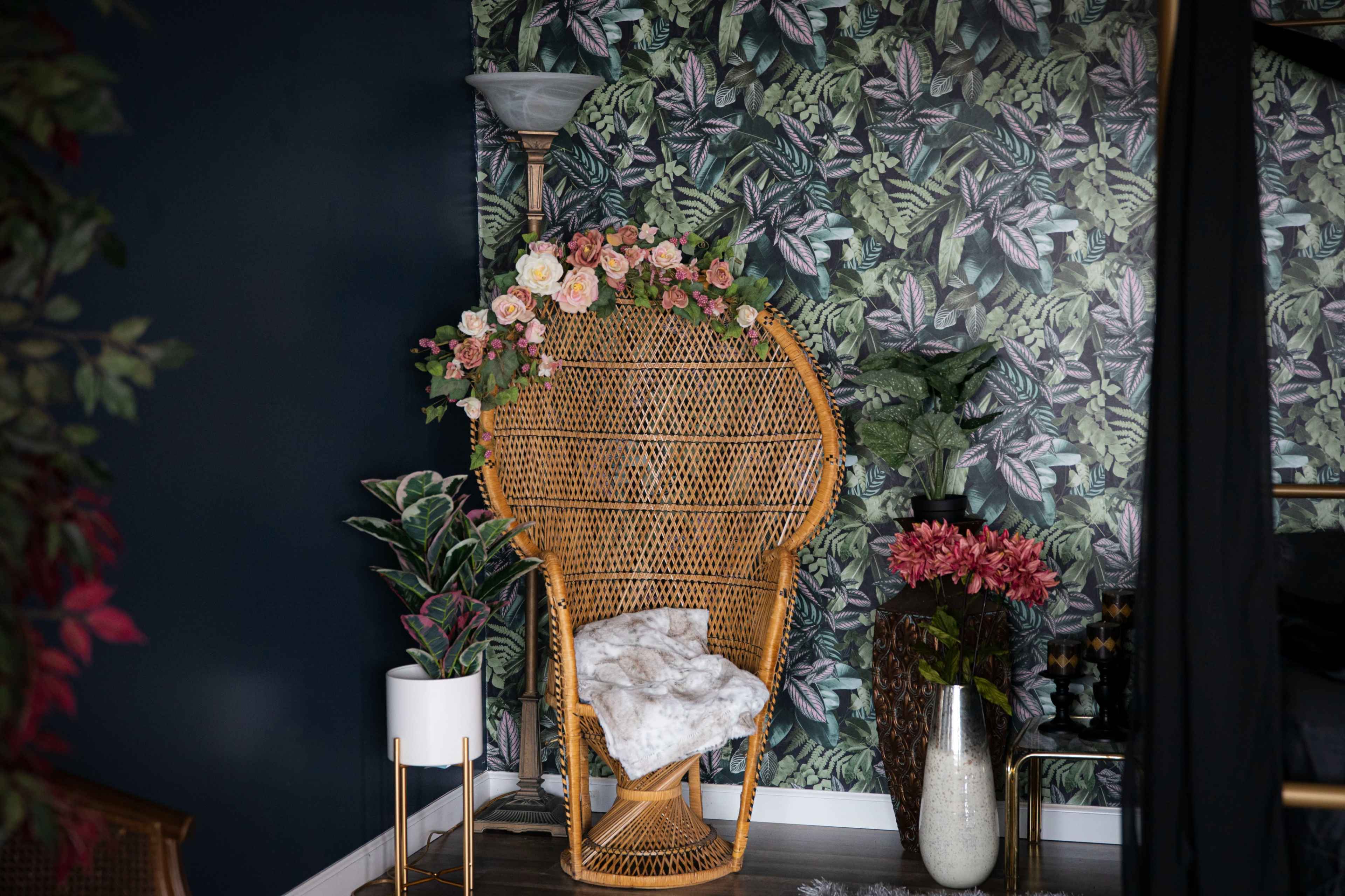 A rattan chair adorned with flowers sits against a wall decorated with tropical-themed wallpaper, surrounded by potted plants.