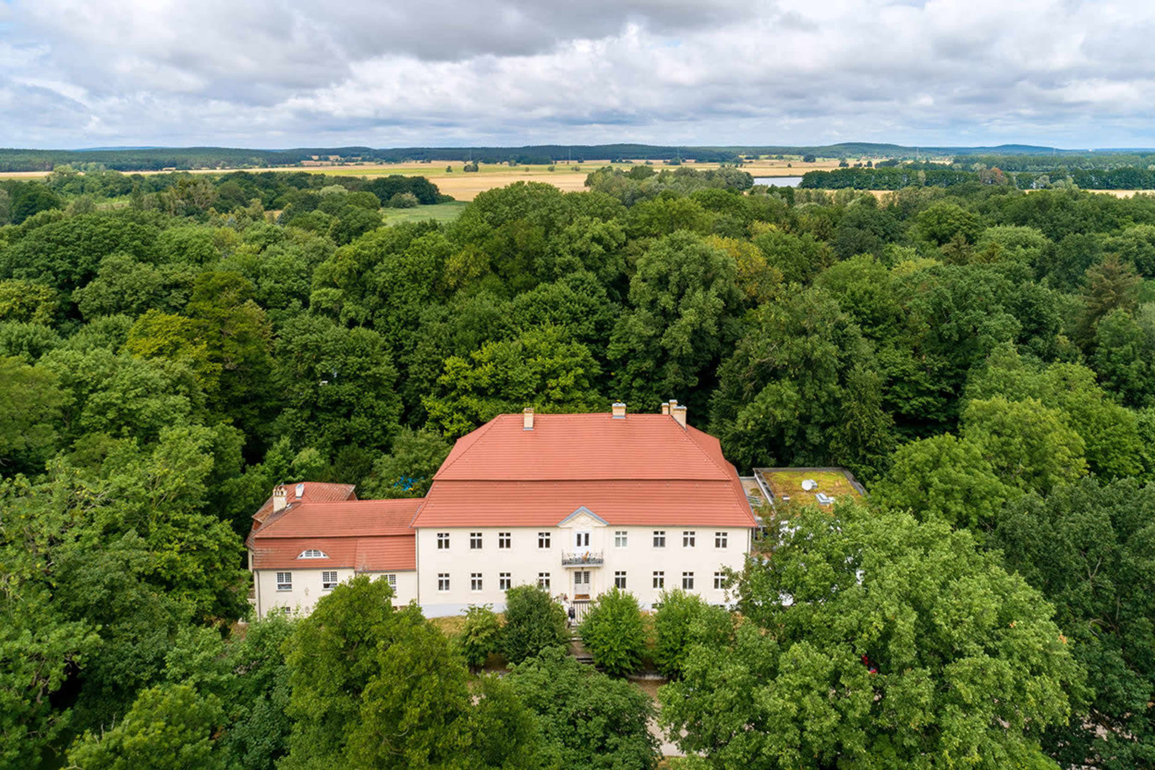 A large white building with a red roof is situated amidst dense green trees.