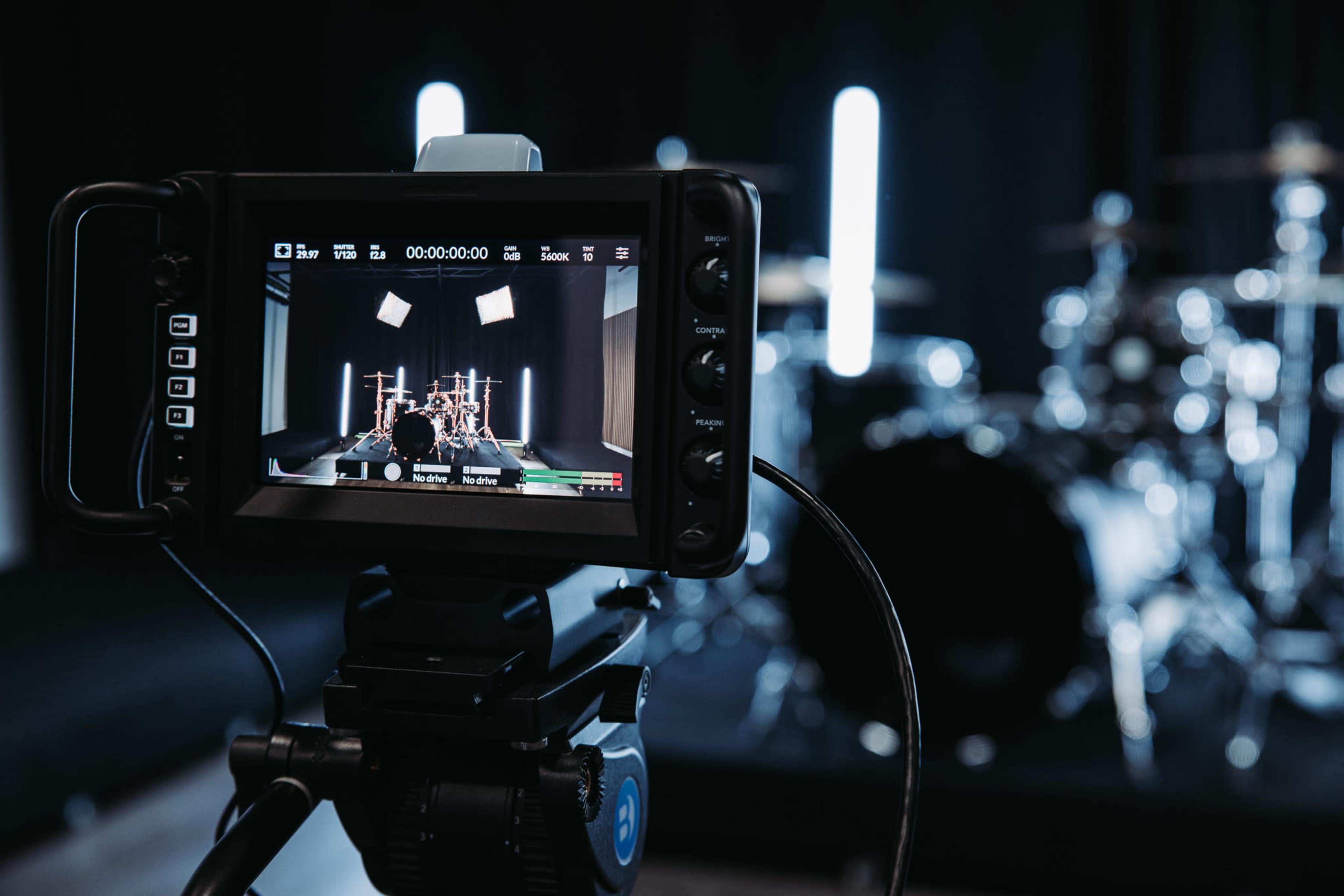 A camera monitor displays a studio setup with a drum kit in the background.