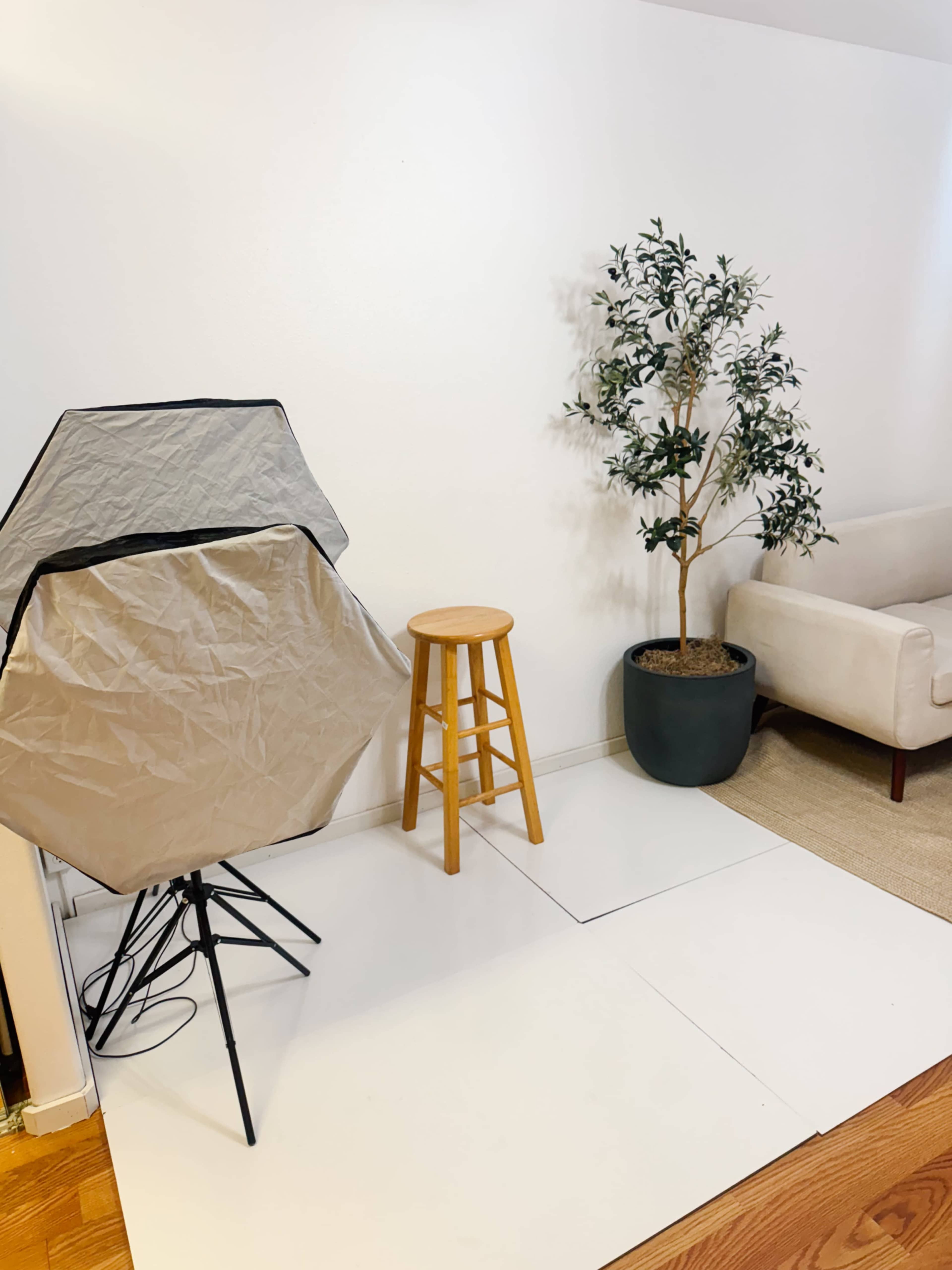 The scene features a photography setup with a light modifier, a wooden stool, a potted plant, and a couch against a white wall.