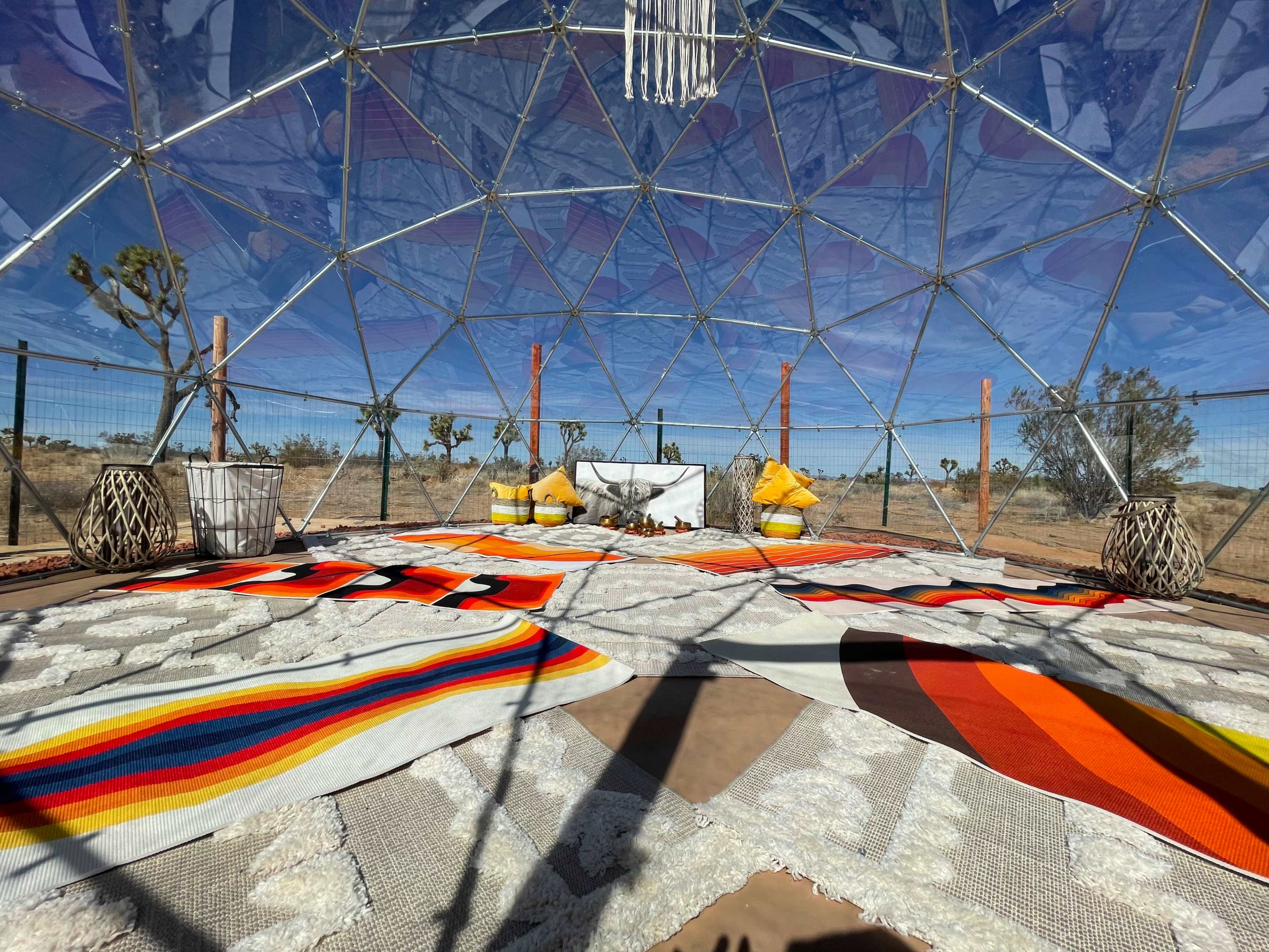 The interior of a geodesic dome features colorful rugs on a sandy floor, with seating arrangements of pillows and a decorative enclave in the center.