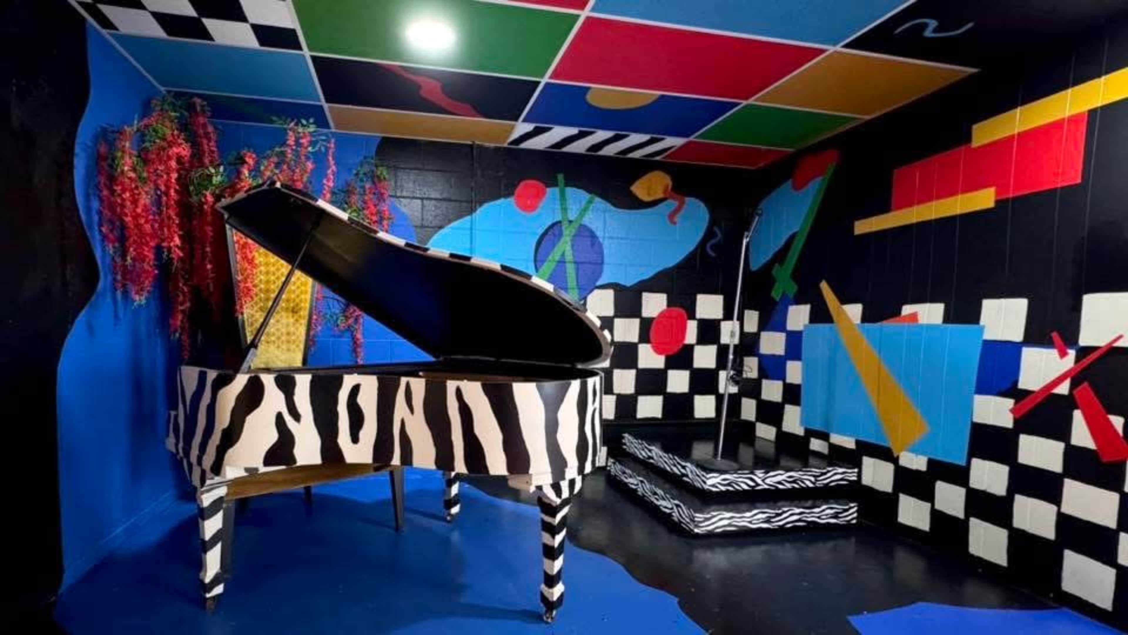 The image shows a room with abstract mural artwork, featuring a zebra-themed grand piano and colorful geometric designs on the walls and ceiling.