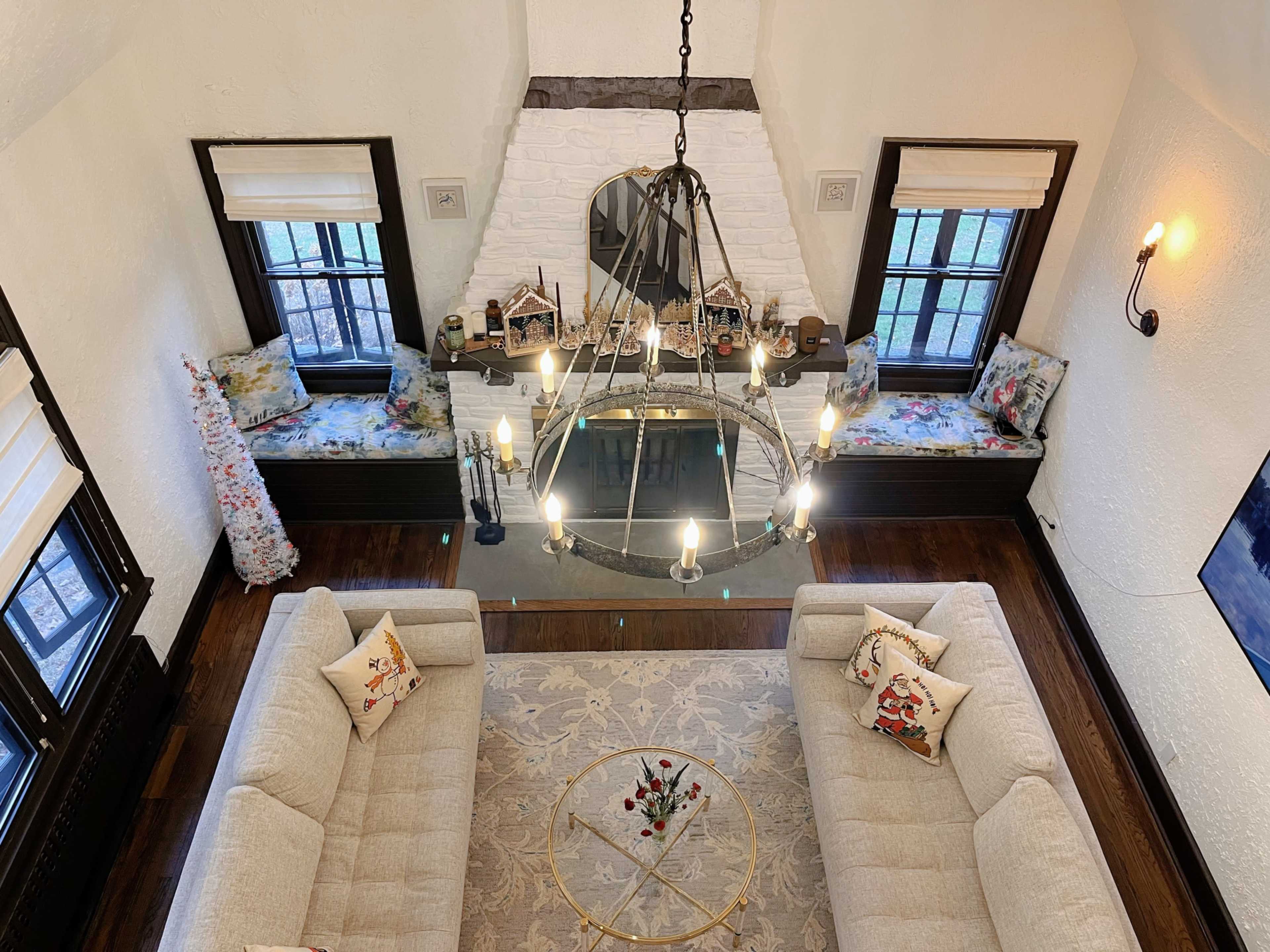 The image shows a living room with a high ceiling, featuring a chandelier, two light-colored sofas, a round coffee table, and a white brick fireplace.