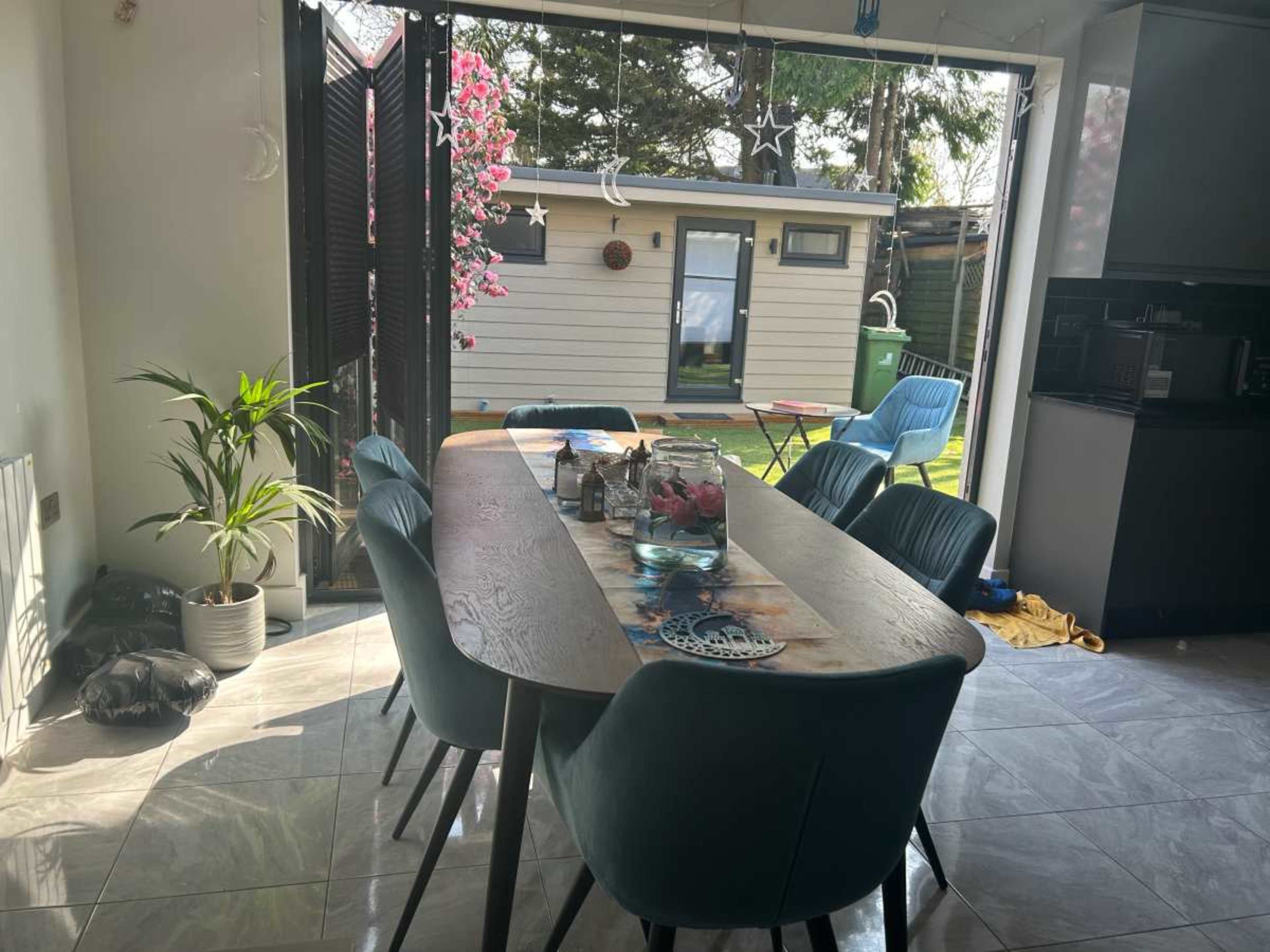 A dining table with blue chairs is set in a brightly lit kitchen, facing an outdoor area featuring a small building and greenery.