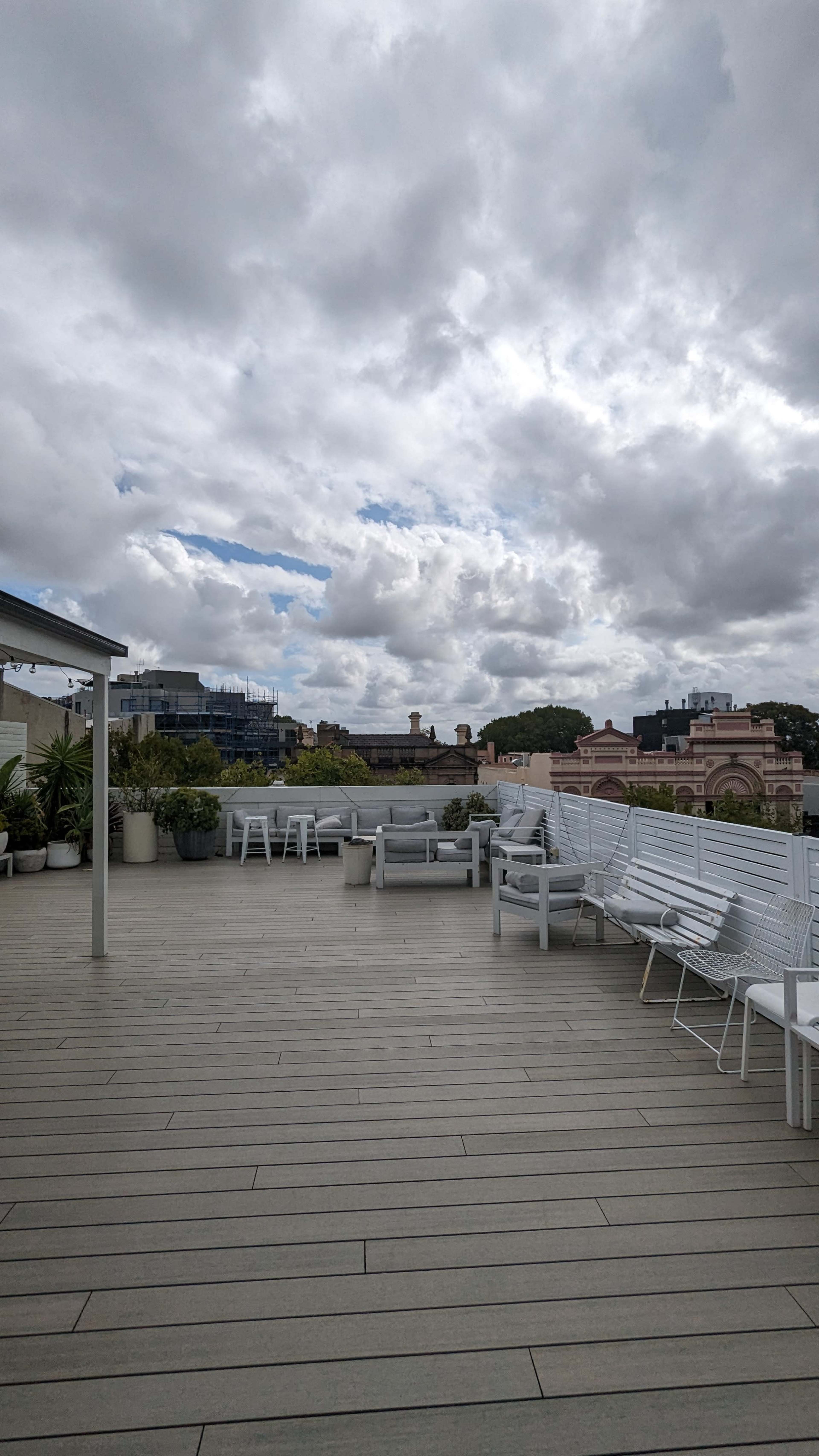 The image shows a rooftop terrace with white chairs and tables, surrounded by cloudy skies and a view of buildings in the background.