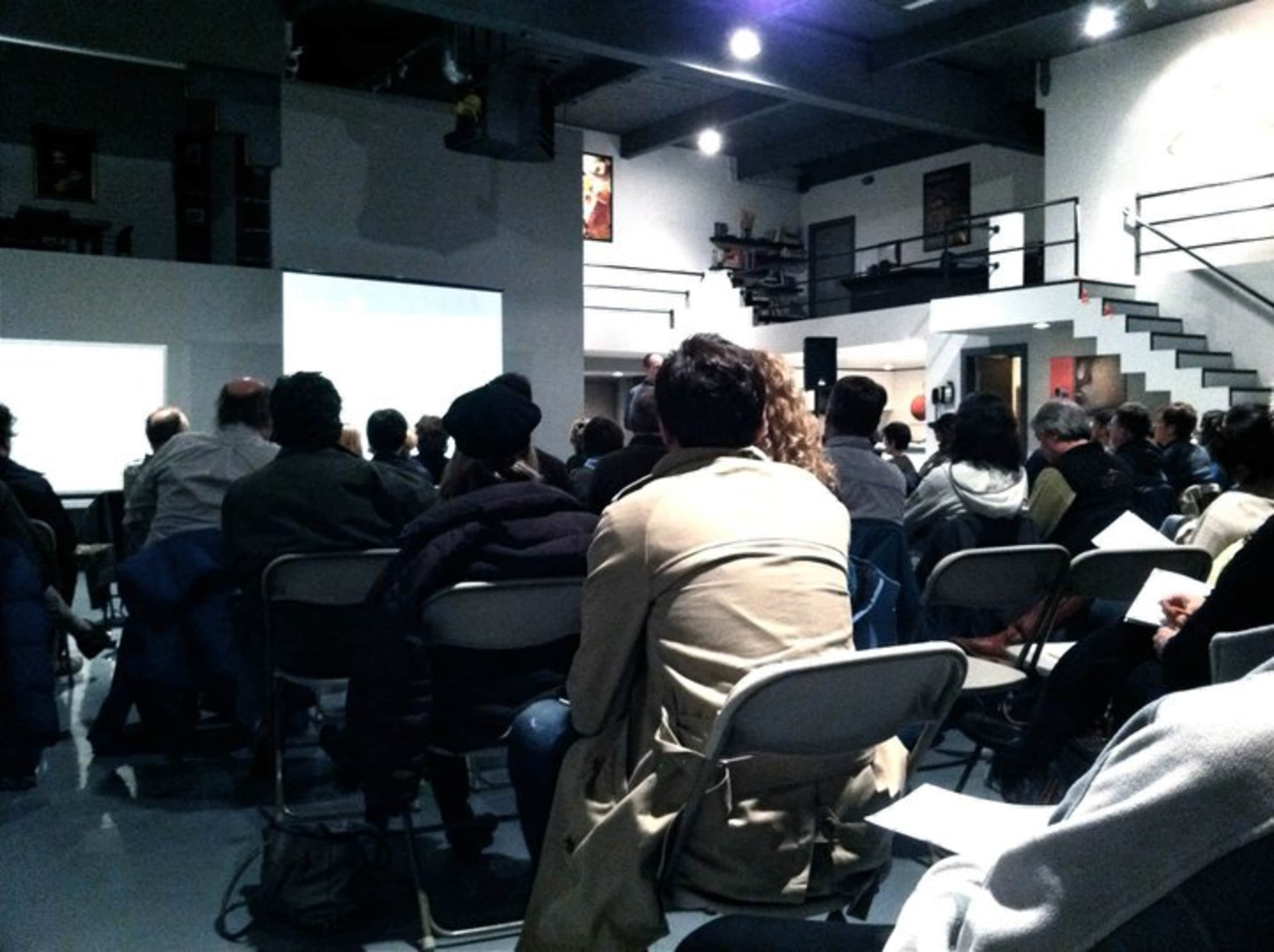 A group of people is seated in a spacious room, watching a presentation on a screen.