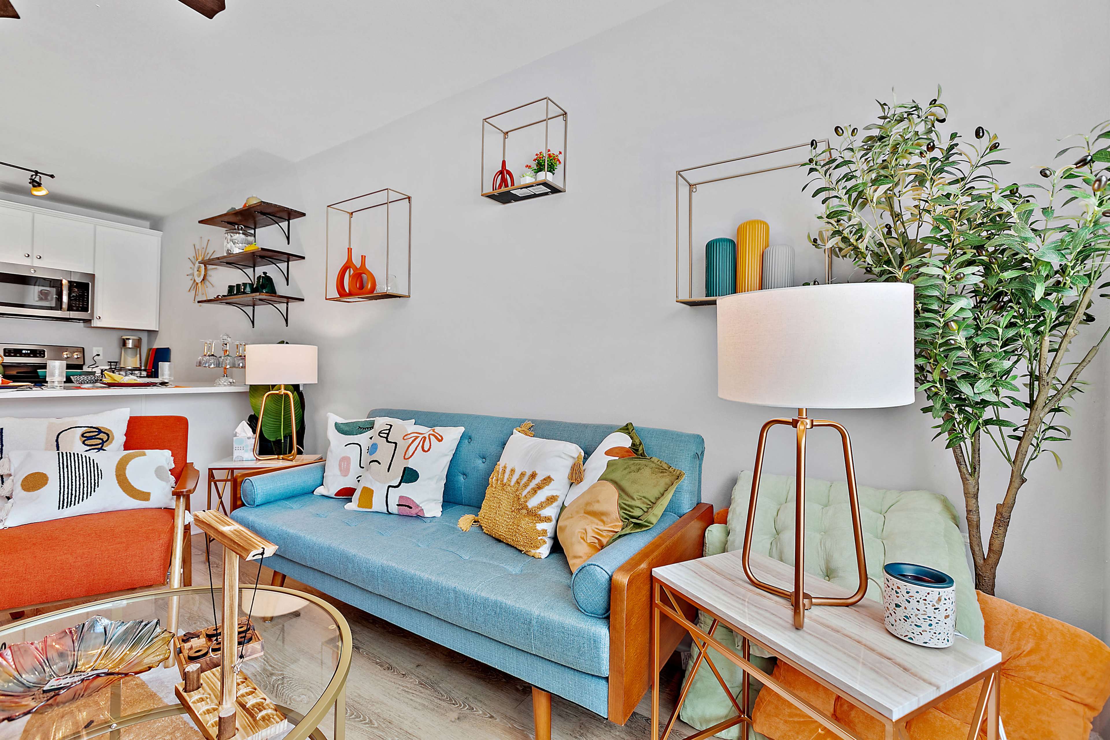 The living room features a blue couch with decorative pillows, an orange armchair, a glass coffee table, and shelves displaying decorative objects against a light gray wall.