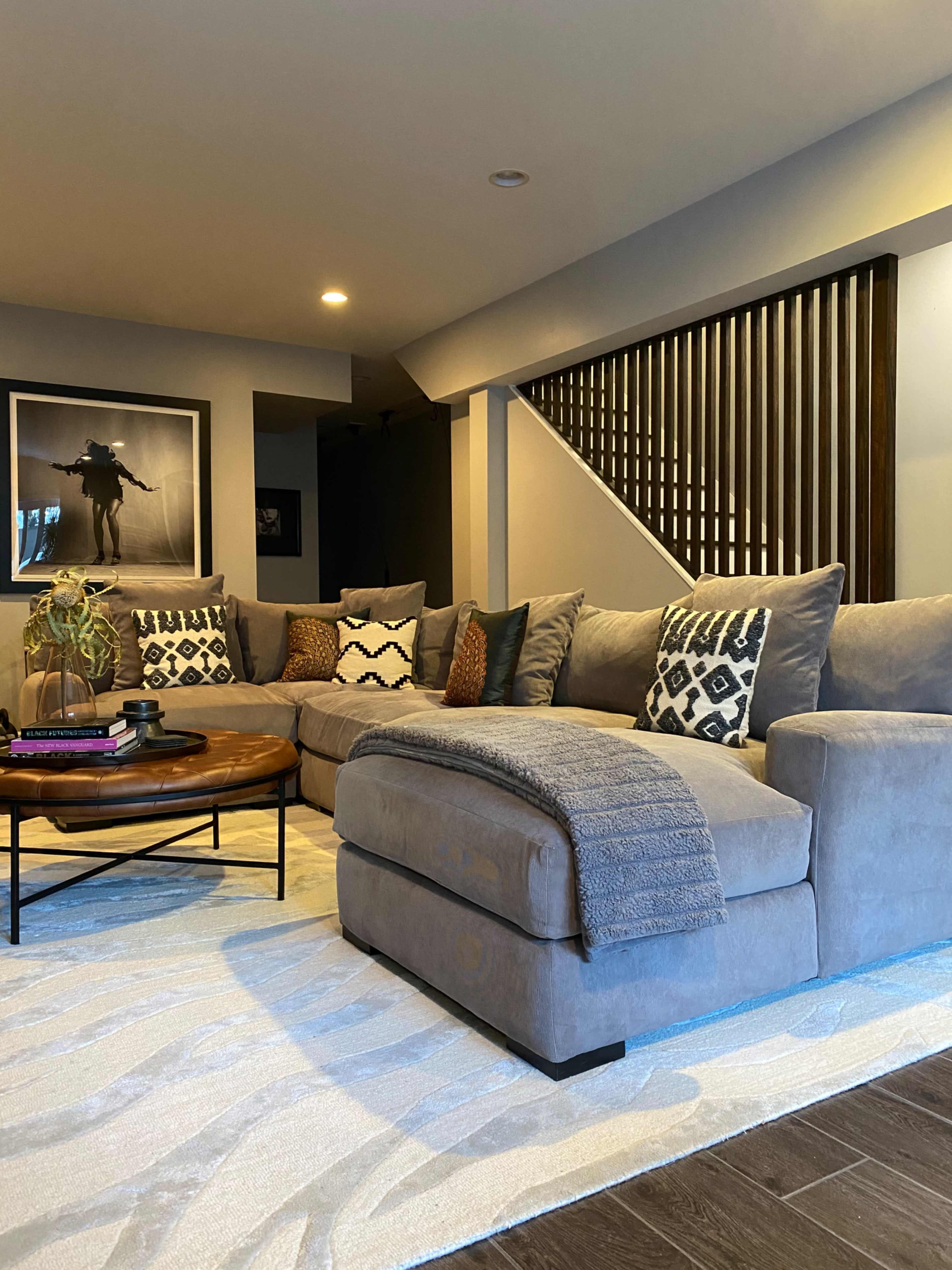 A cozy living room features a gray sectional sofa with patterned pillows, a circular wooden coffee table, and a cream-colored area rug.