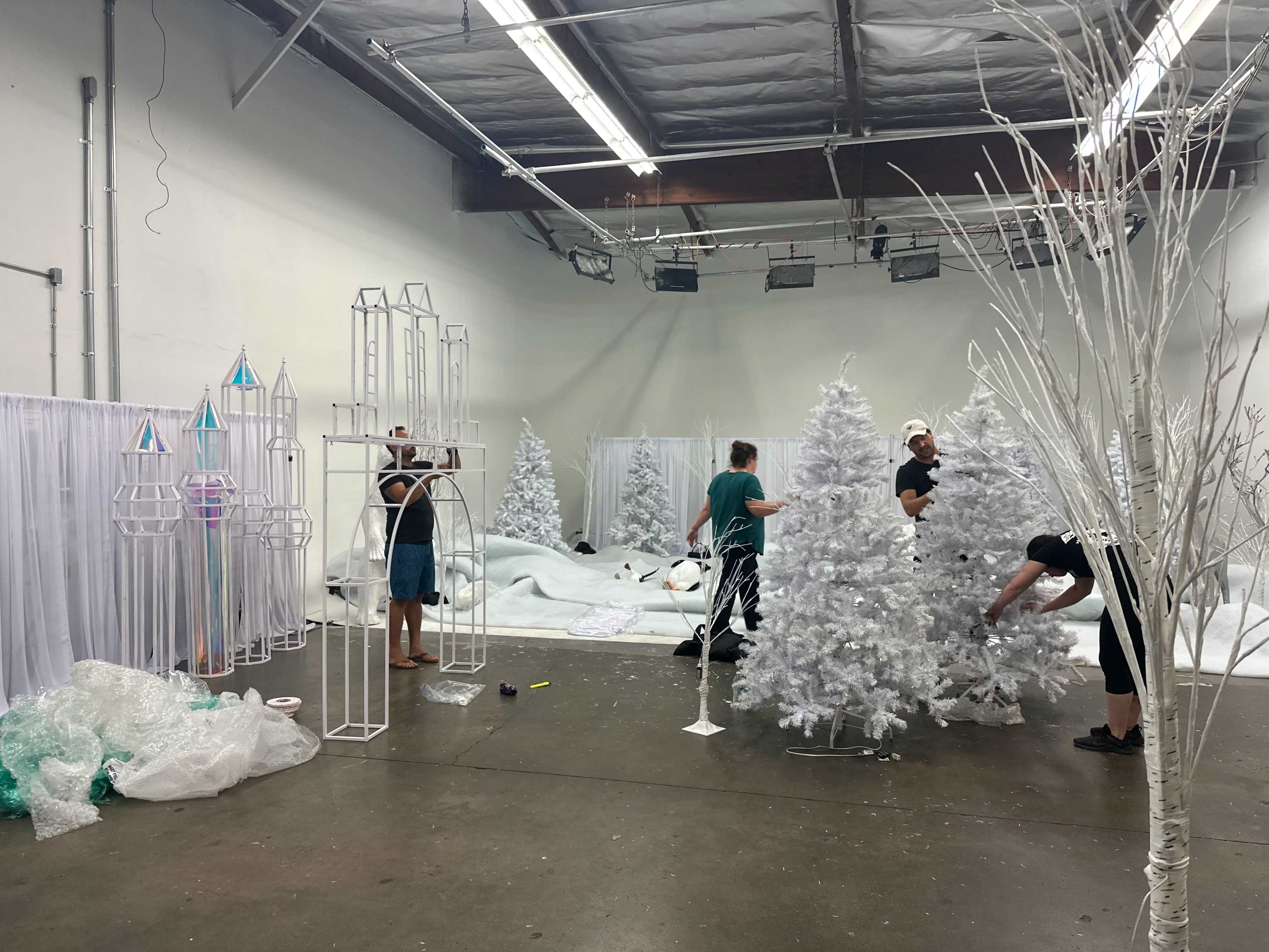 A group of people is assembling white Christmas trees and decorative structures in a large, empty studio space.