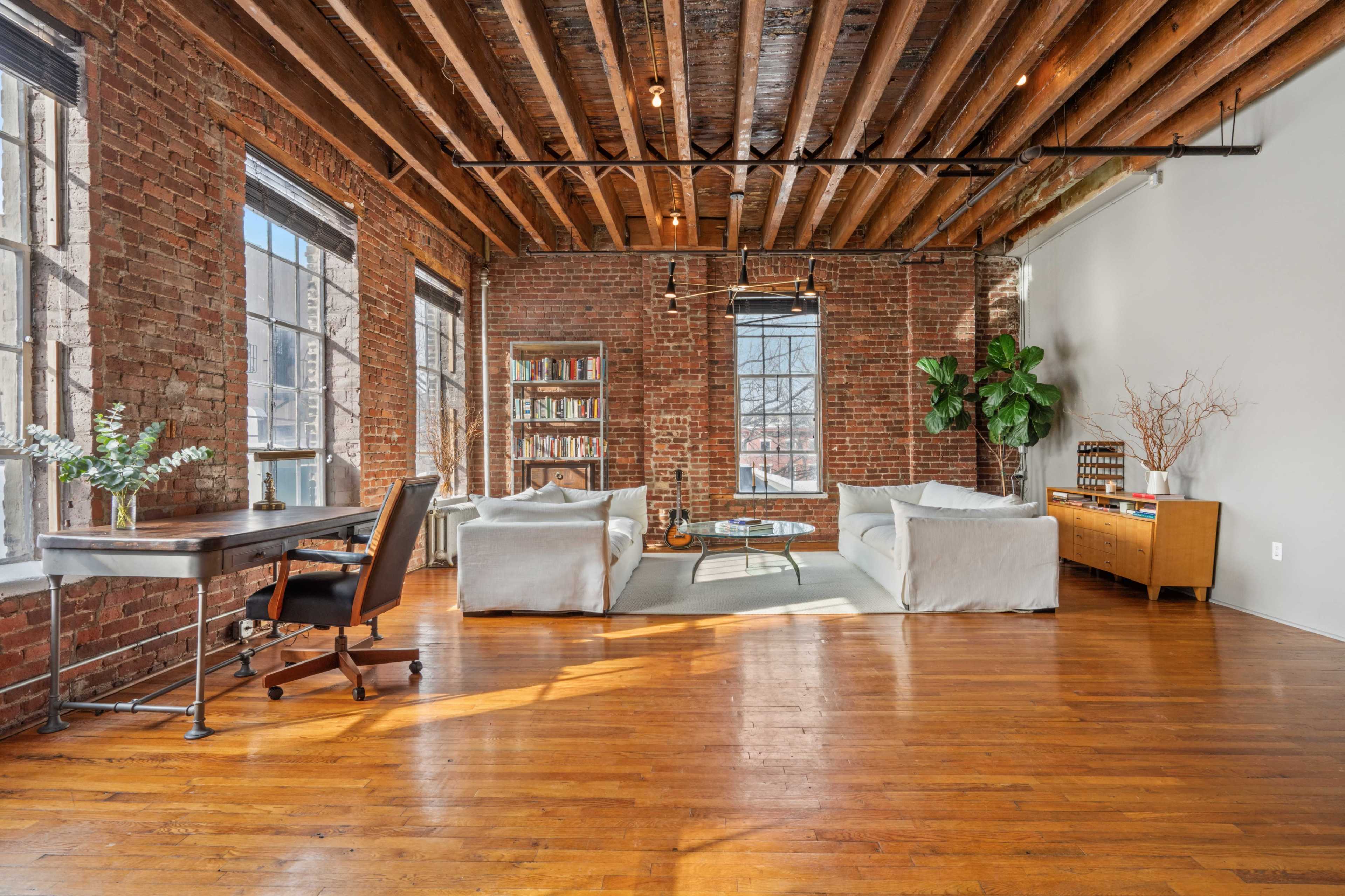 The image shows a spacious living area featuring exposed brick walls, wooden beams, and large windows, with a desk, two sofas, a coffee table, and a bookshelf.