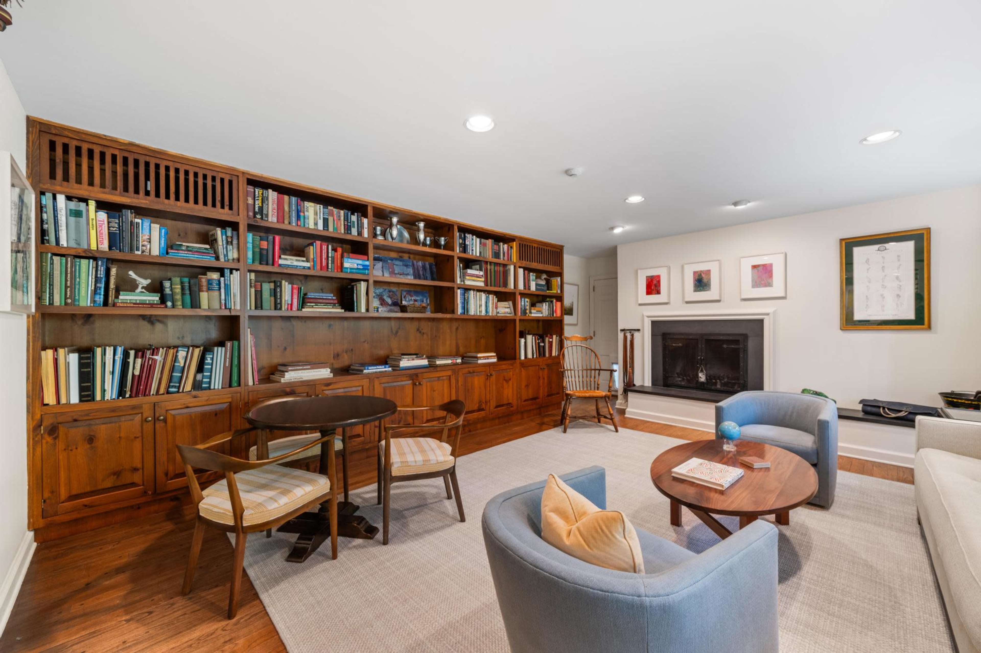 The image shows a cozy living room with a wooden bookshelf filled with books, a fireplace, and two circular tables surrounded by upholstered chairs.