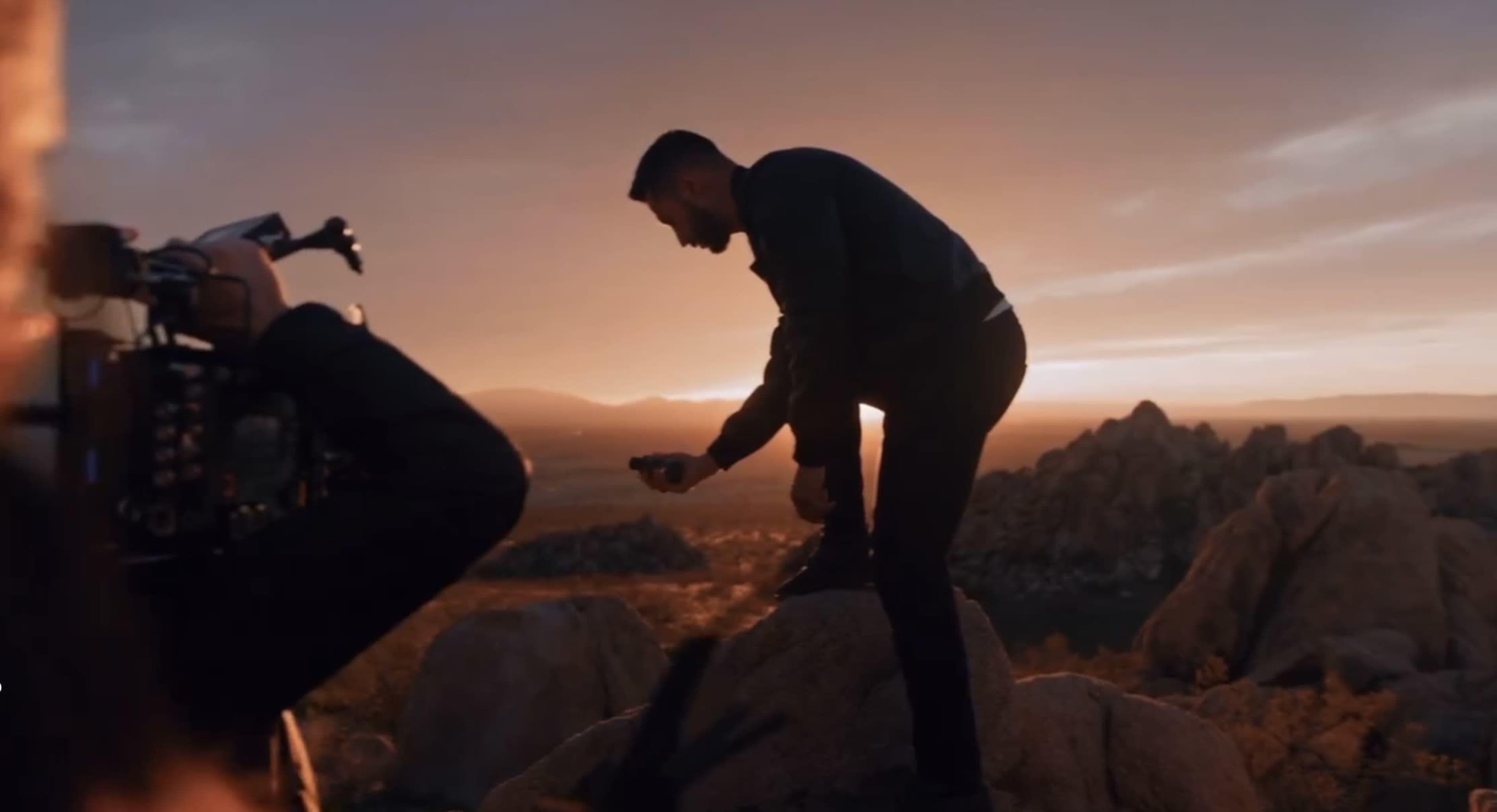 A person stands on a rock at sunset, holding a small object while another person films them with a camera.