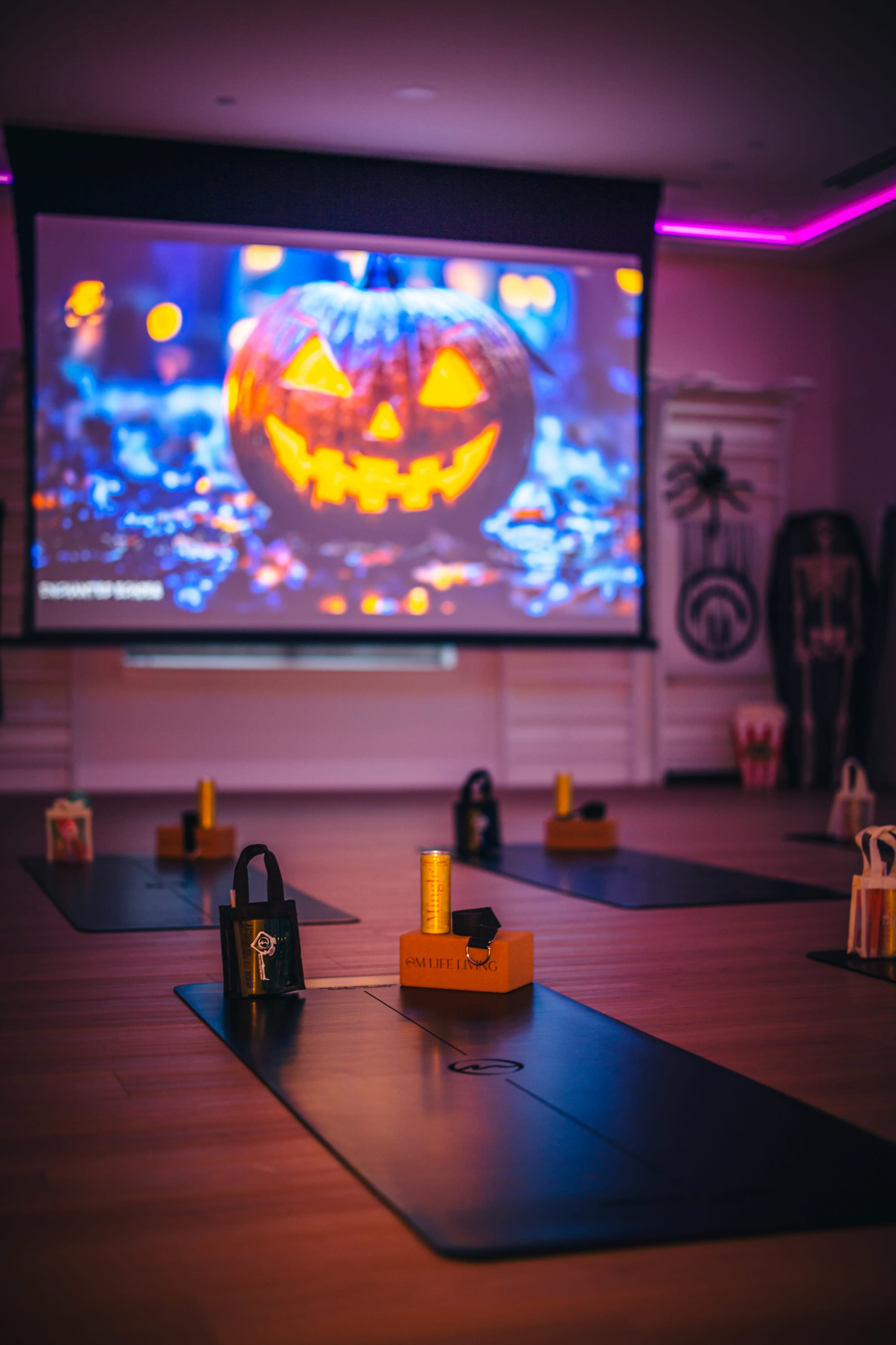 The image shows a yoga studio set up for a Halloween-themed event, featuring yoga mats with decorative items and a large screen displaying a glowing jack-o'-lantern.