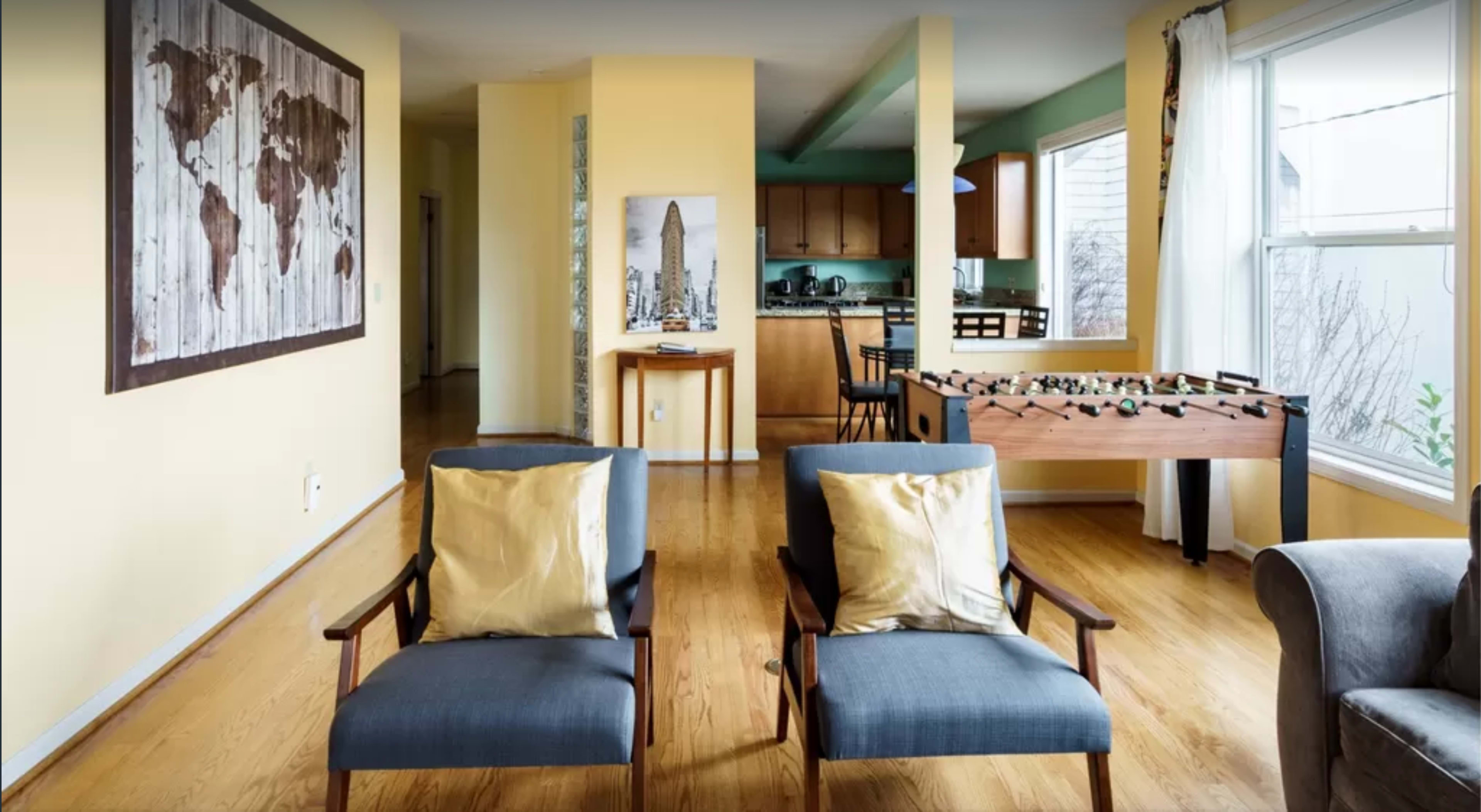 A living room with two chairs facing a wooden table, a foosball table, and a kitchen area in the background.