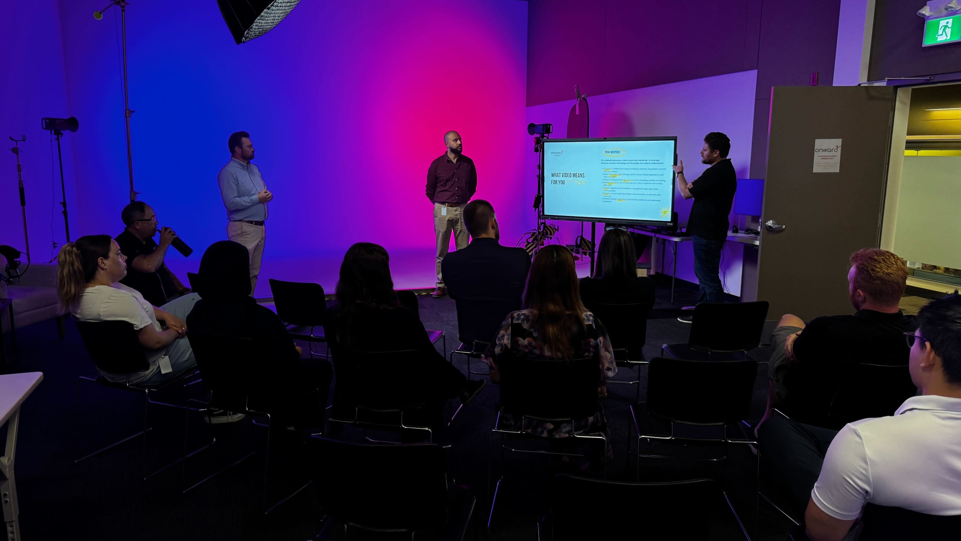 A group of people is seated in chairs, watching a presentation on a screen, while two presenters stand in front against a brightly lit background.