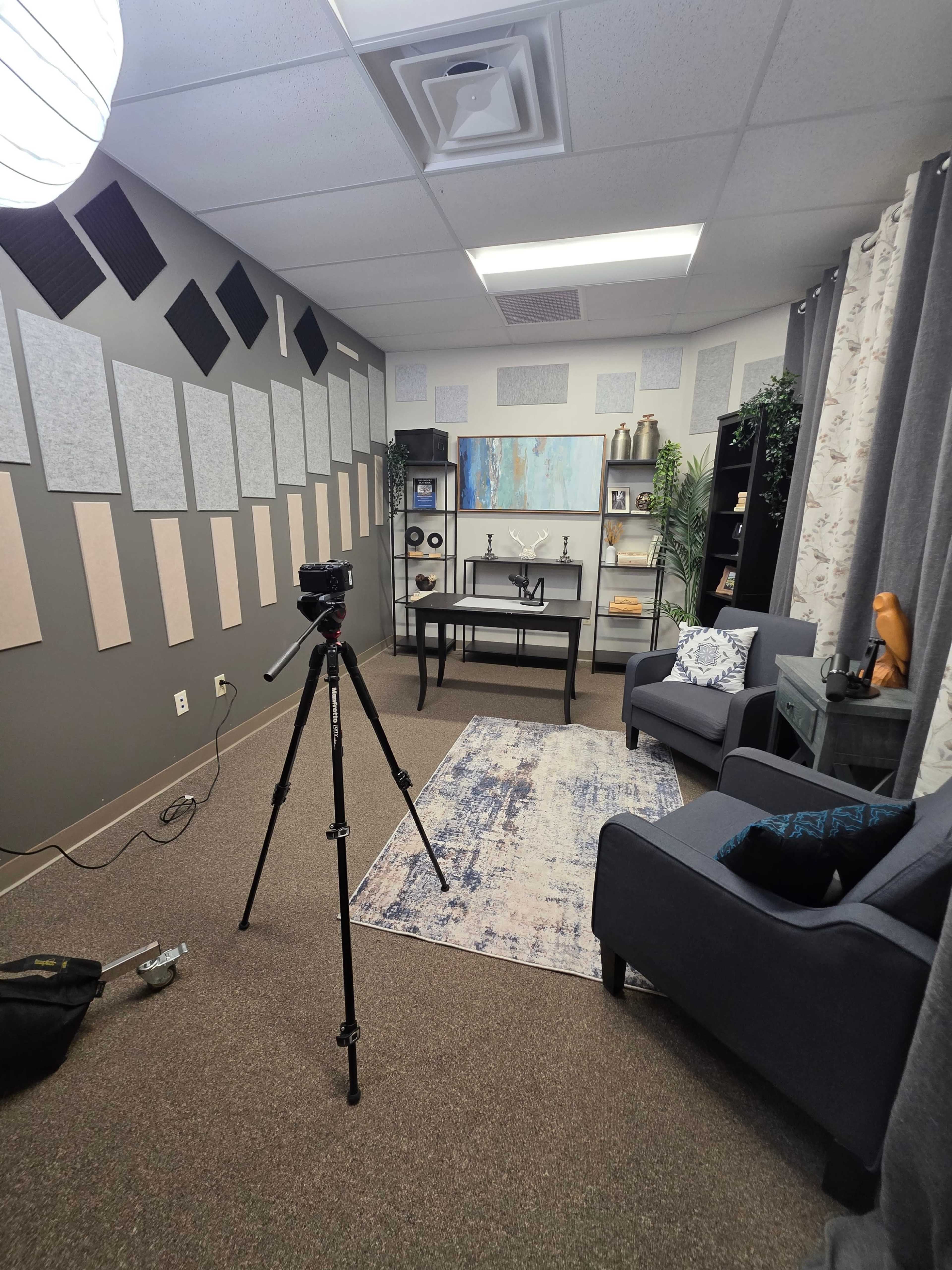 The image shows a small, neatly arranged room with a gray carpet, a desk, two chairs, a camera on a tripod, and soundproofing panels on the walls.