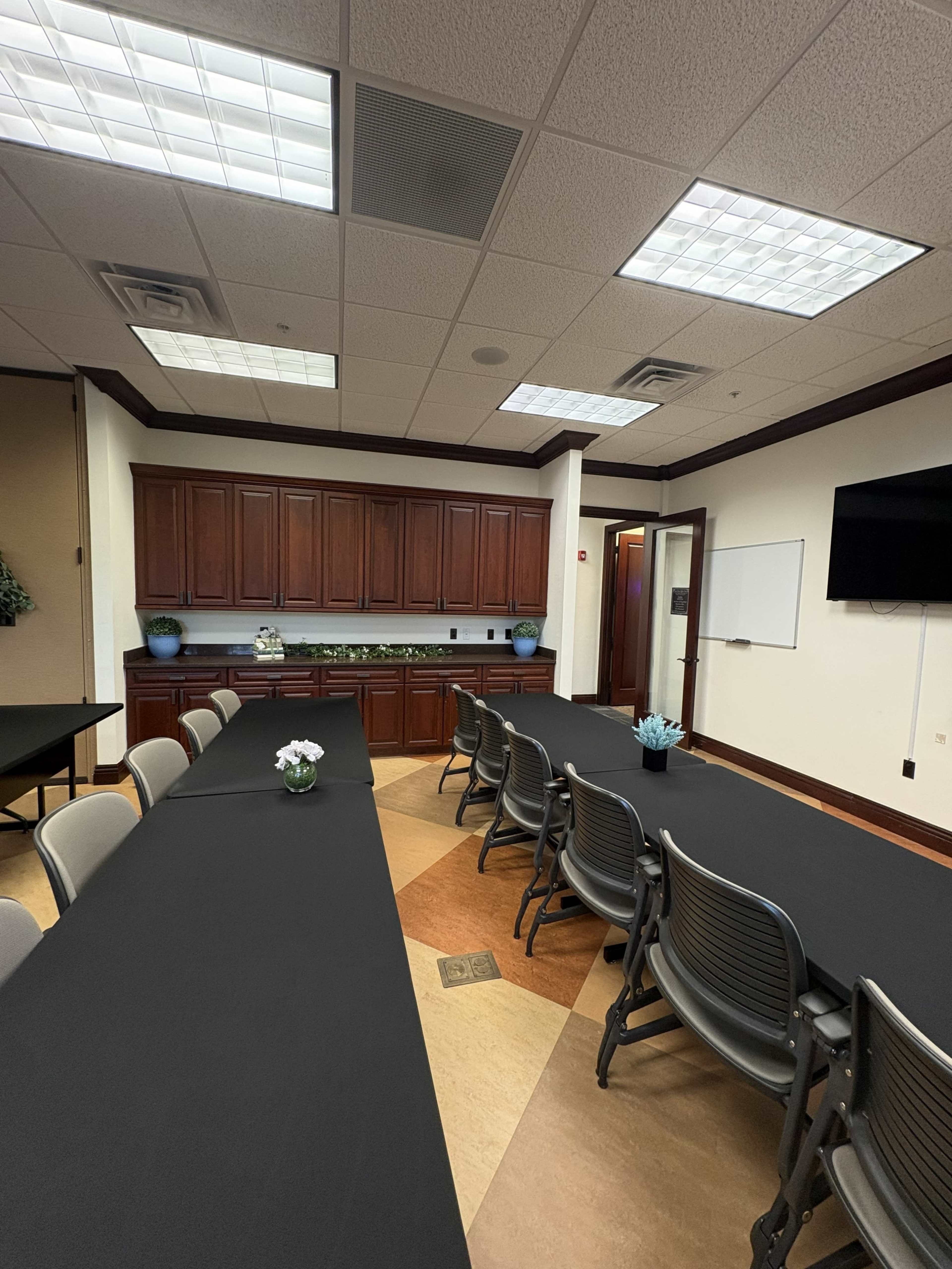 The image shows a conference room with two long black tables arranged in a U-shape and wooden cabinets along the back wall.