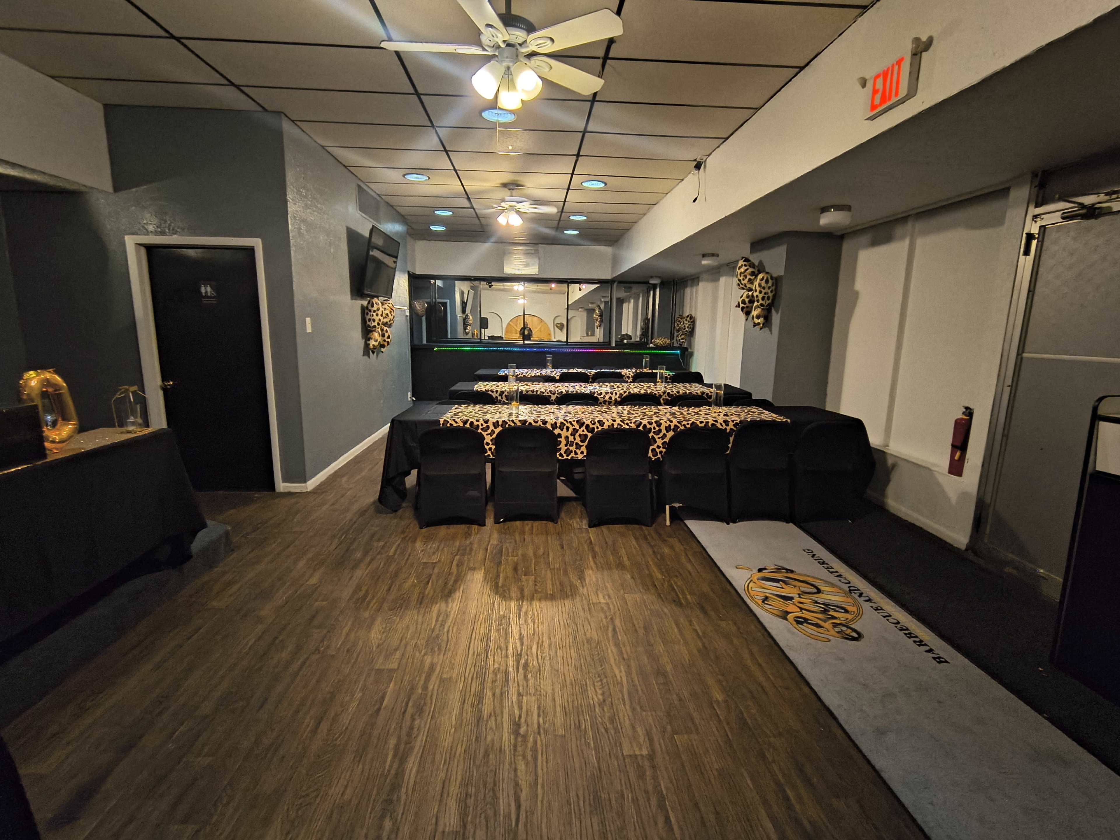 The image shows a spacious event room with a long table covered in a leopard-print tablecloth and black chairs, along with a bar area and a television on the wall.