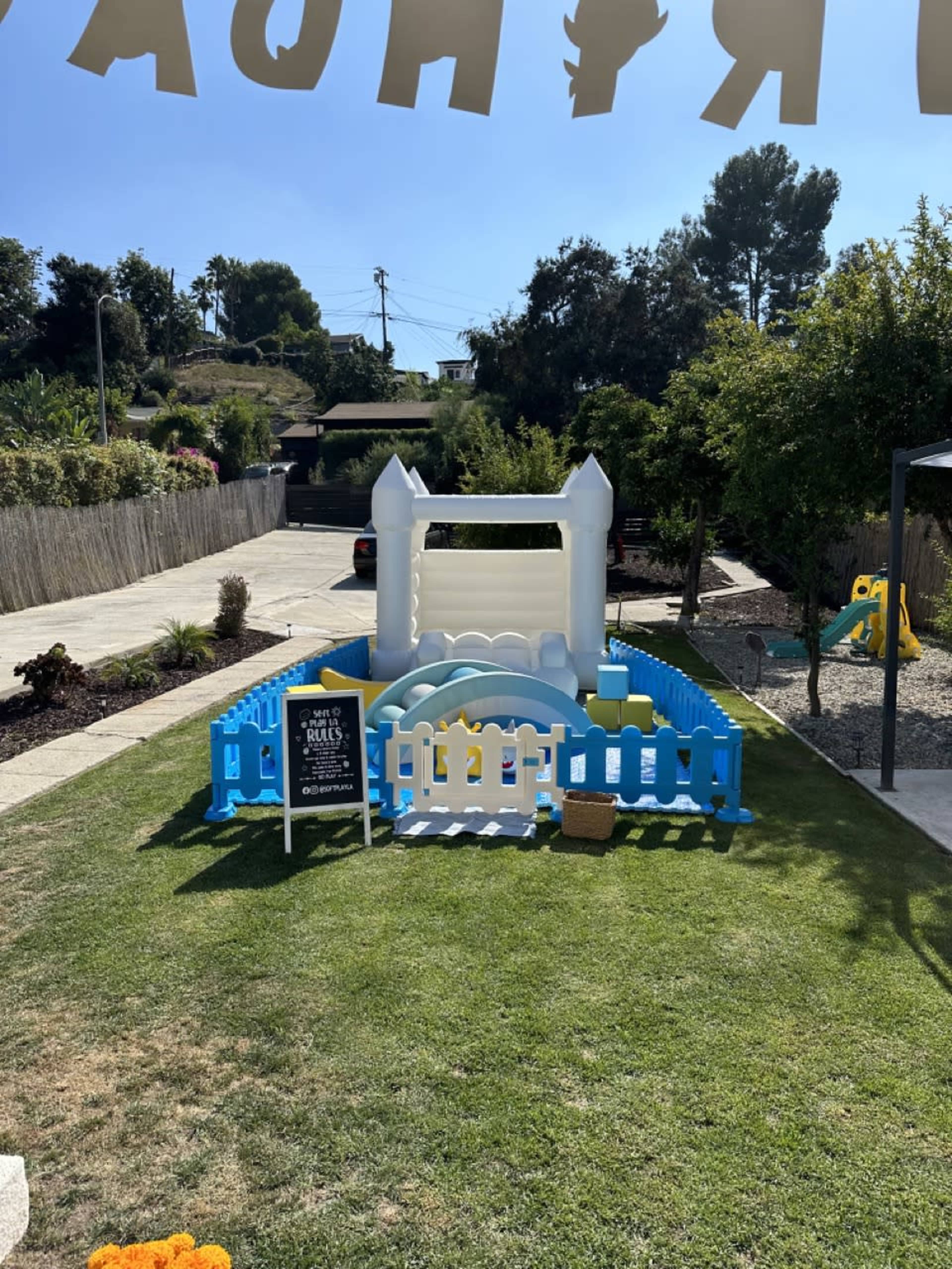 The image shows a colorful inflatable bounce house enclosed by a blue and white fence in a grassy yard with trees in the background.