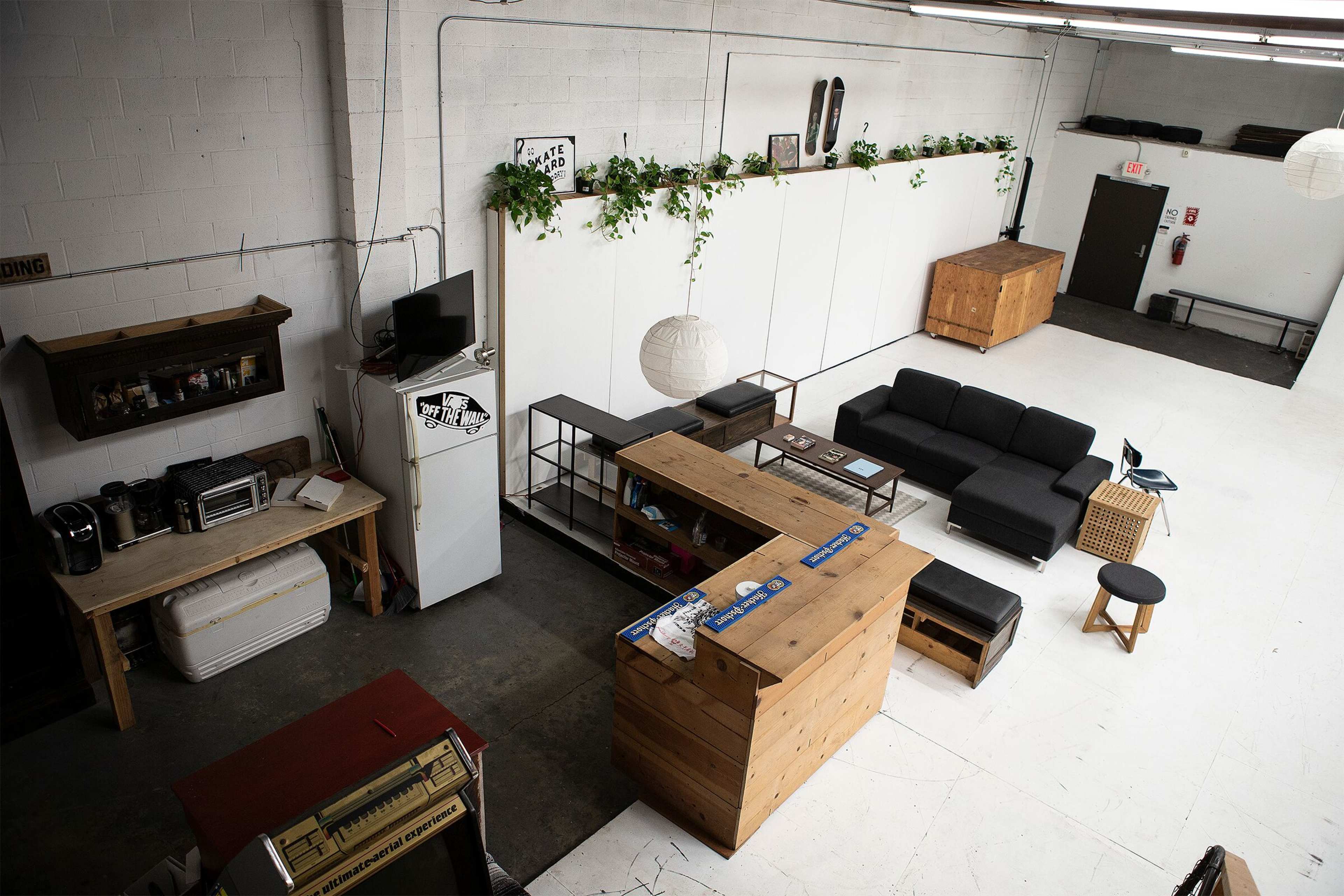 A spacious, minimalist interior with a black sofa, wooden furniture, a kitchenette area, and plants along the walls.