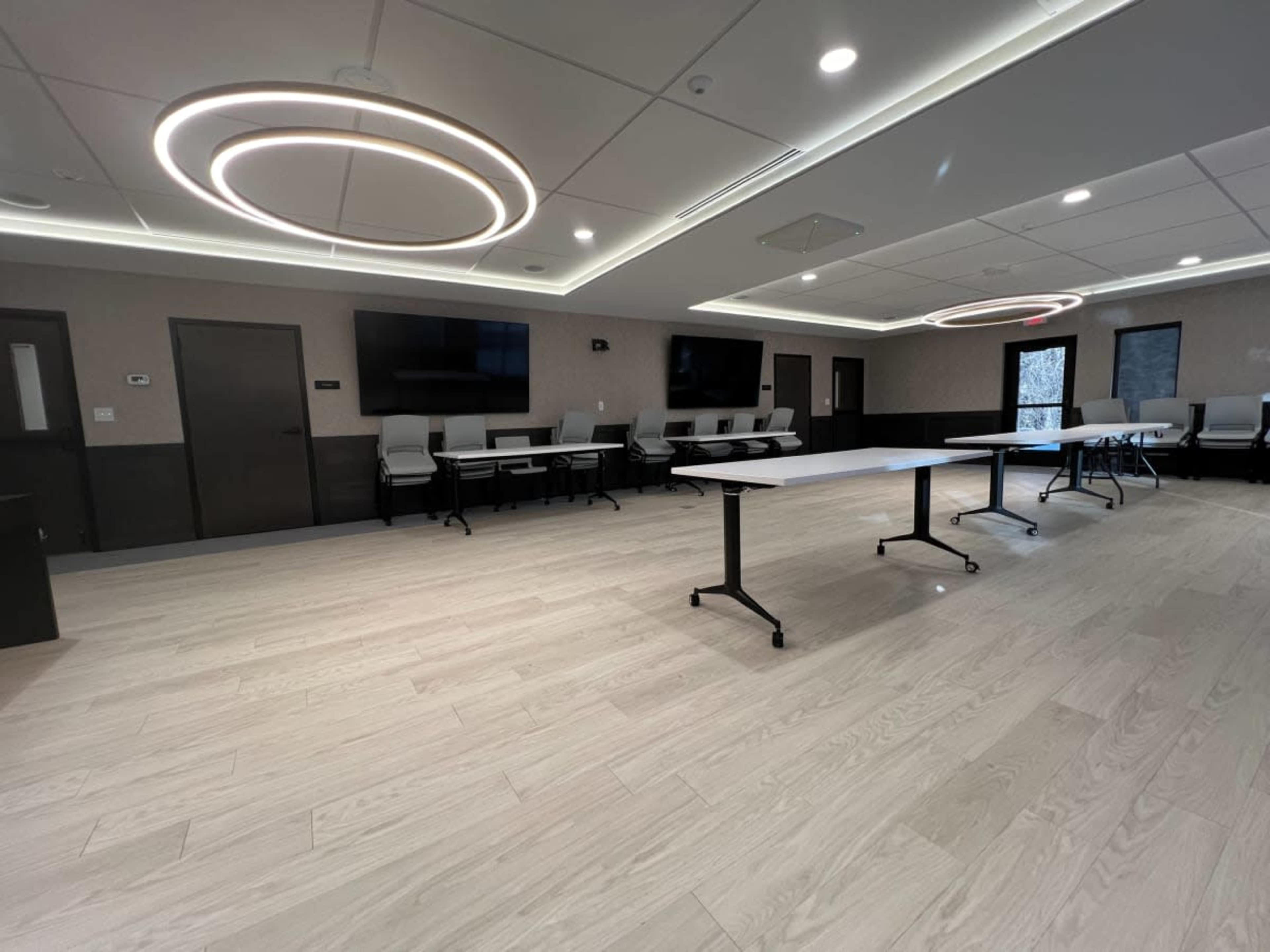 A modern meeting room features two circular ceiling lights, multiple flat-screen monitors, and movable tables arranged on a light wooden floor.