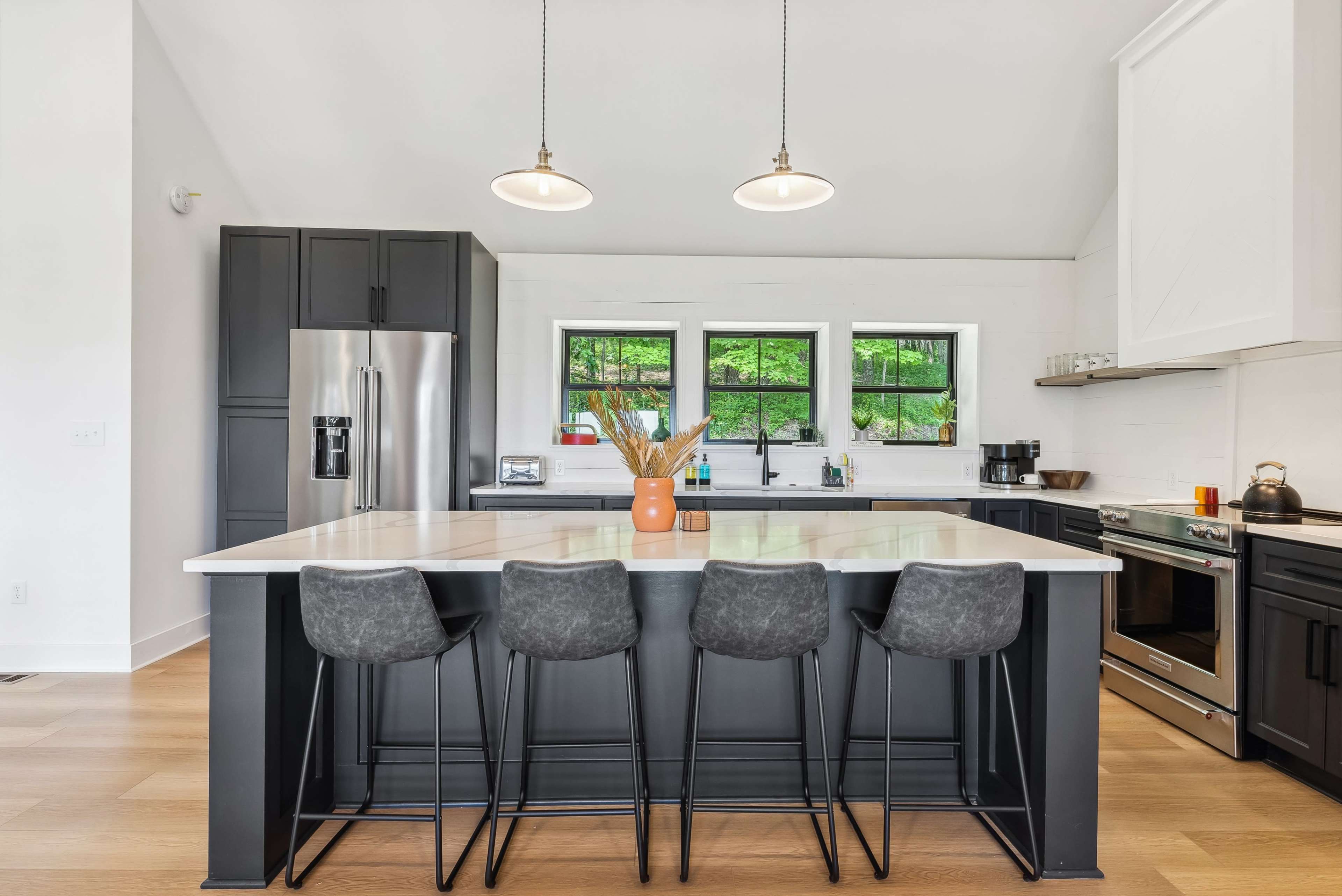 A modern kitchen features a large island with four black bar stools and stainless steel appliances against a backdrop of white cabinetry and green outdoor views.