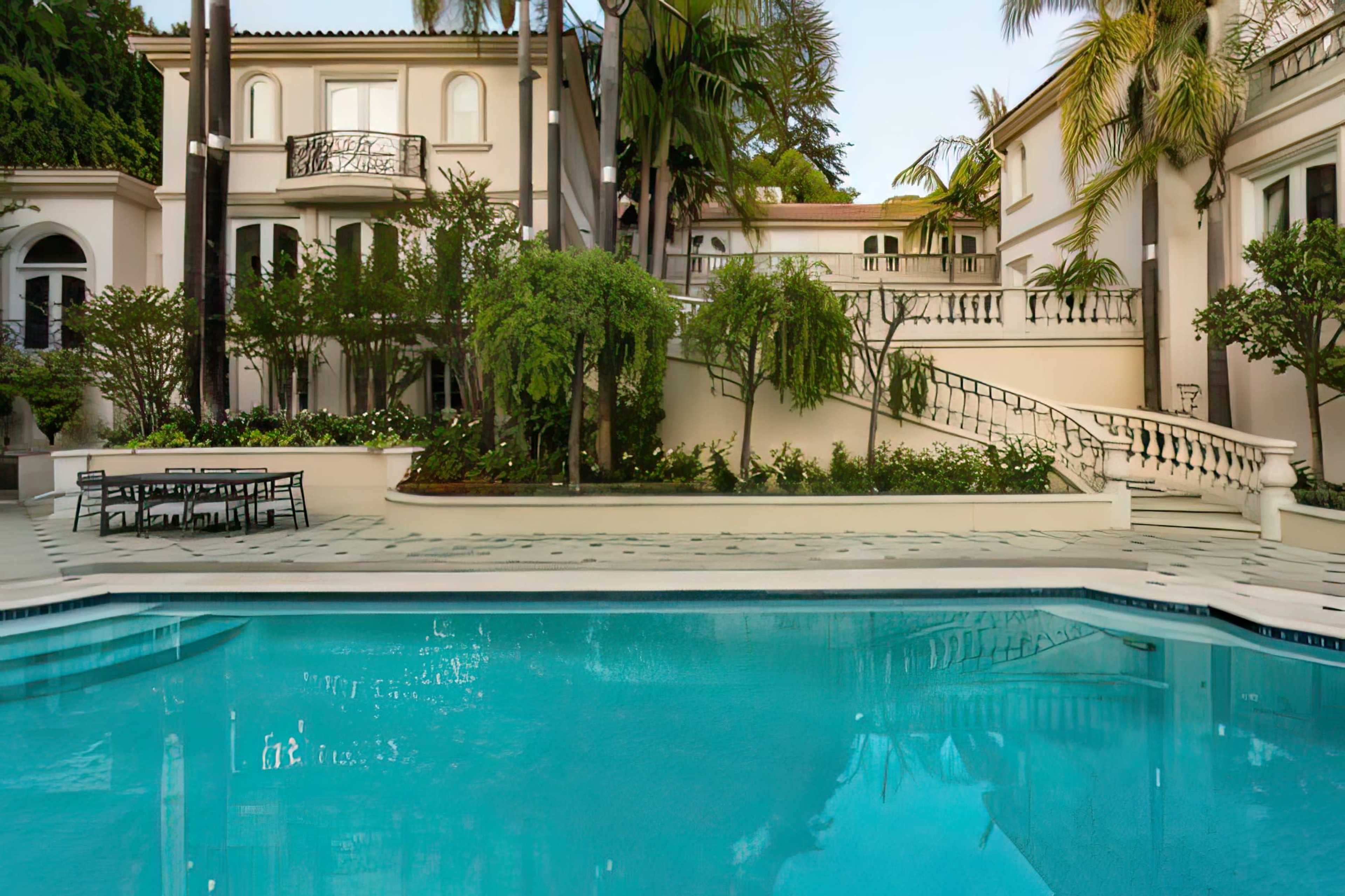 The image shows a luxurious pool in a landscaped backyard of a grand house, surrounded by palm trees and elegant architecture.