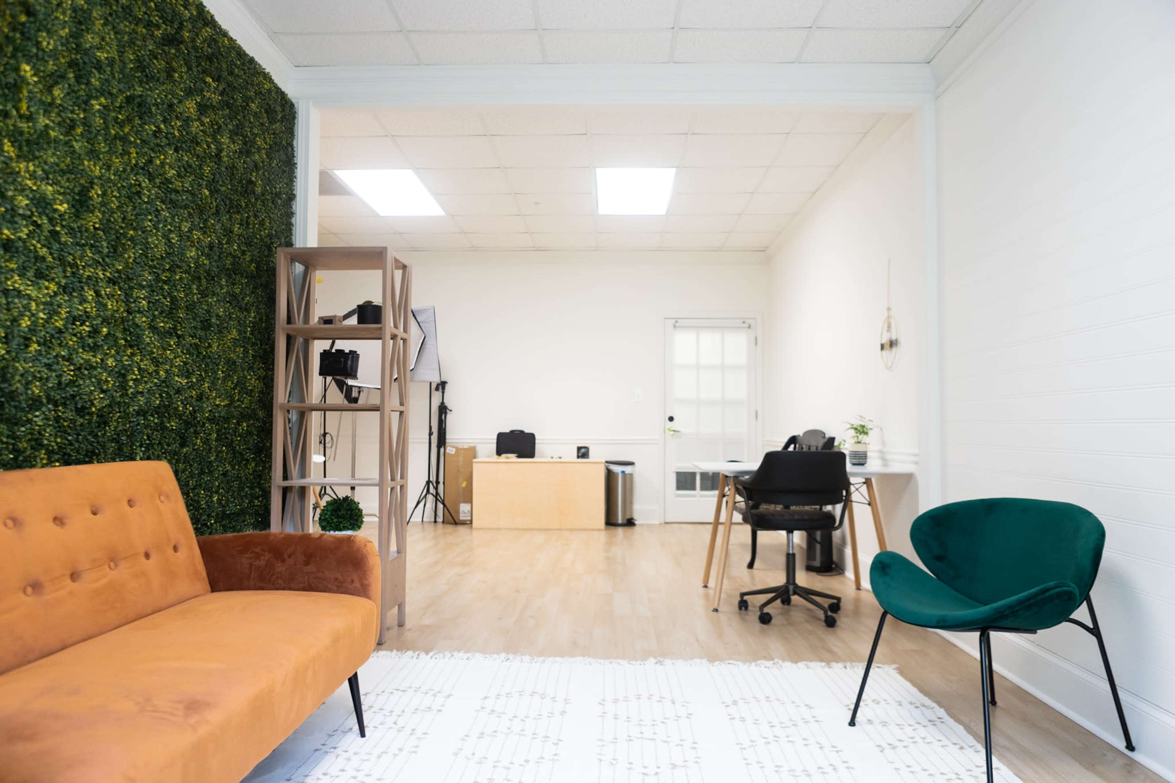 The image shows a modern office space with a green wall, an orange sofa, a wooden shelf, and a workspace featuring a desk and a black chair.