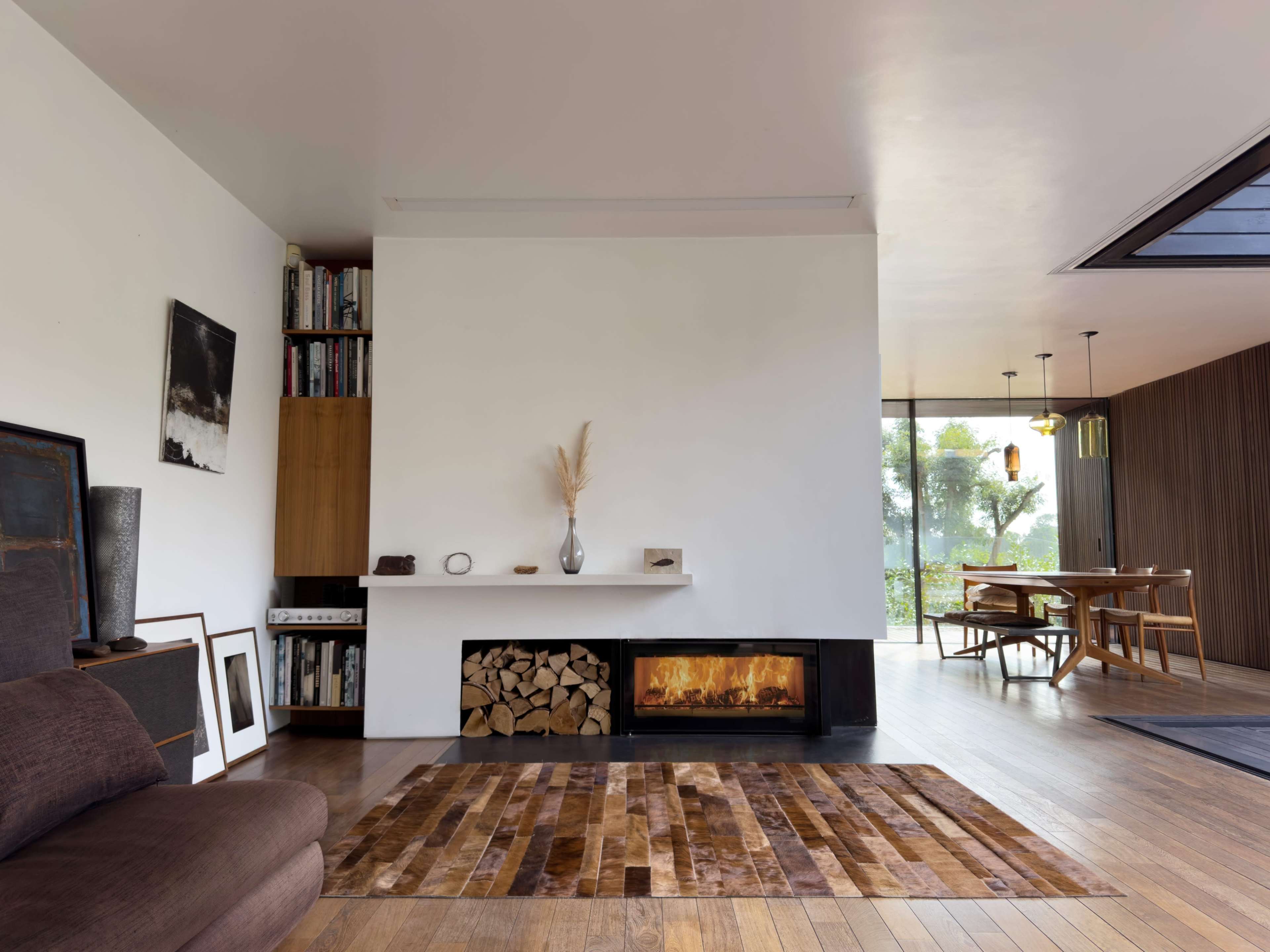 The image shows a modern living room with a minimalist design, featuring a cozy seating area, a fireplace stacked with firewood, and a dining area with wooden furniture.