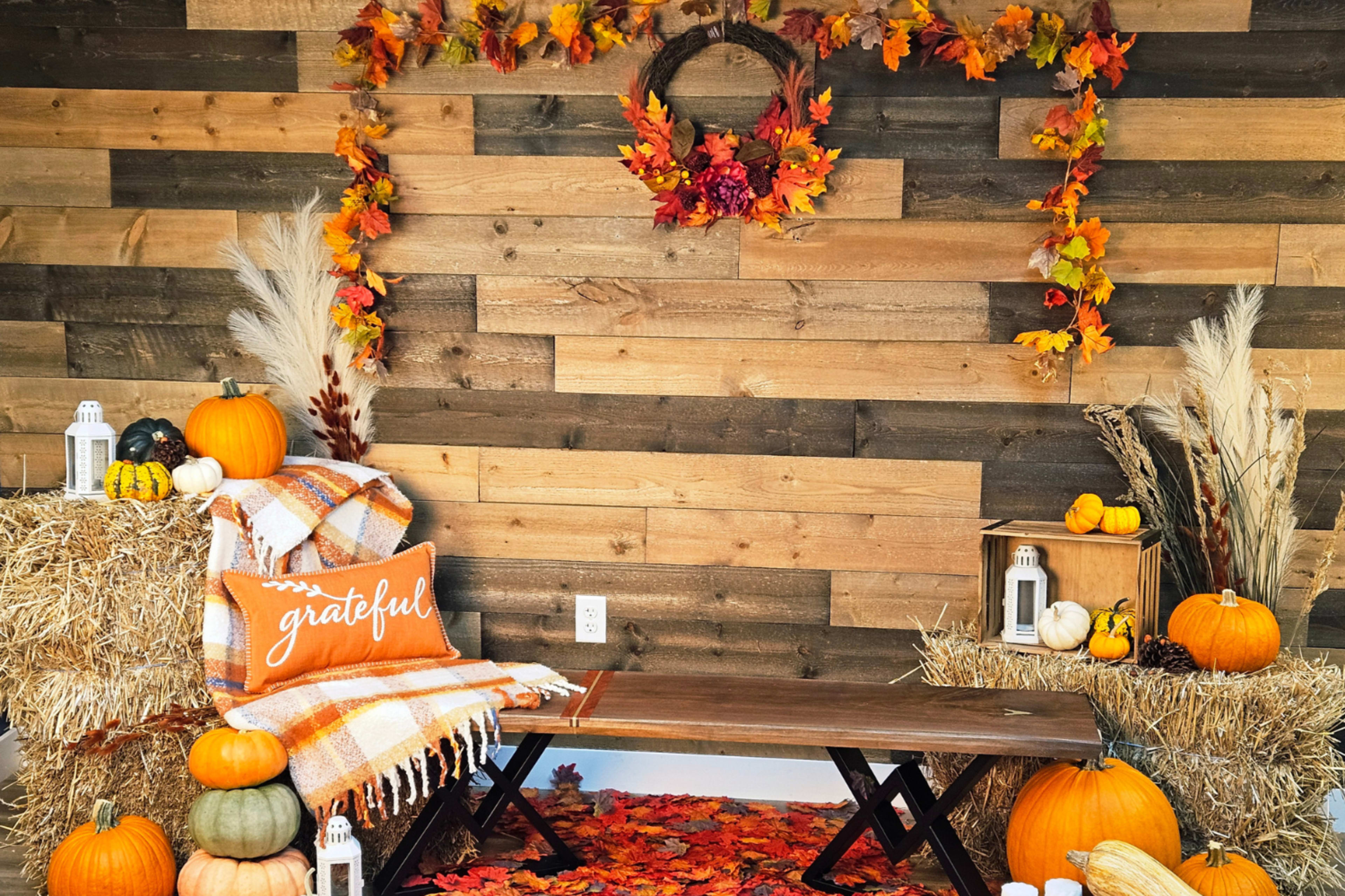 The image features a rustic fall-themed display with pumpkins, straw bales, and autumn leaves arranged around a wooden bench adorned with a blanket and a "grateful" pillow.