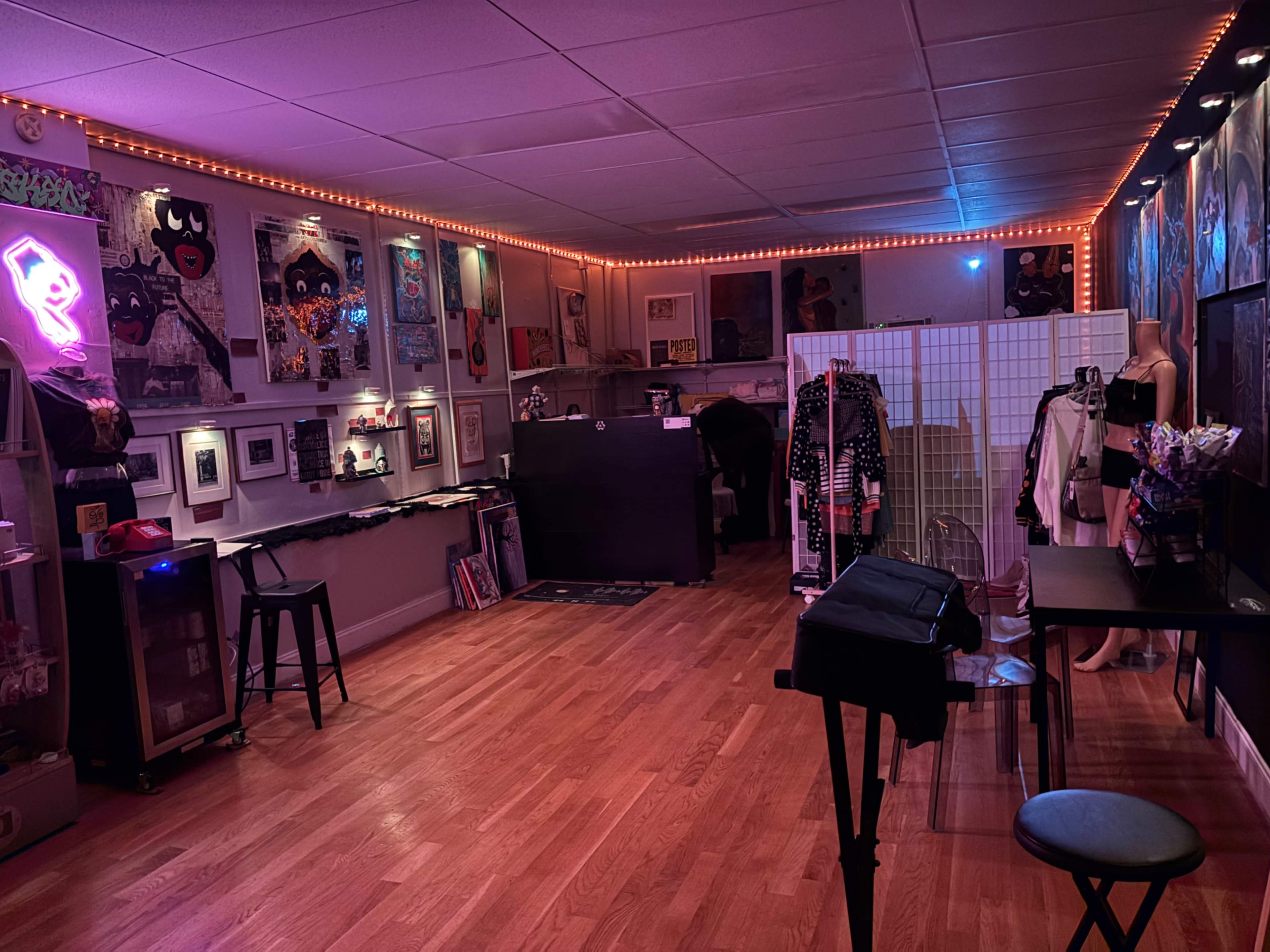 The image shows a brightly lit art studio or gallery featuring colorful artwork on the walls, wooden flooring, and various furniture items including tables and display stands.