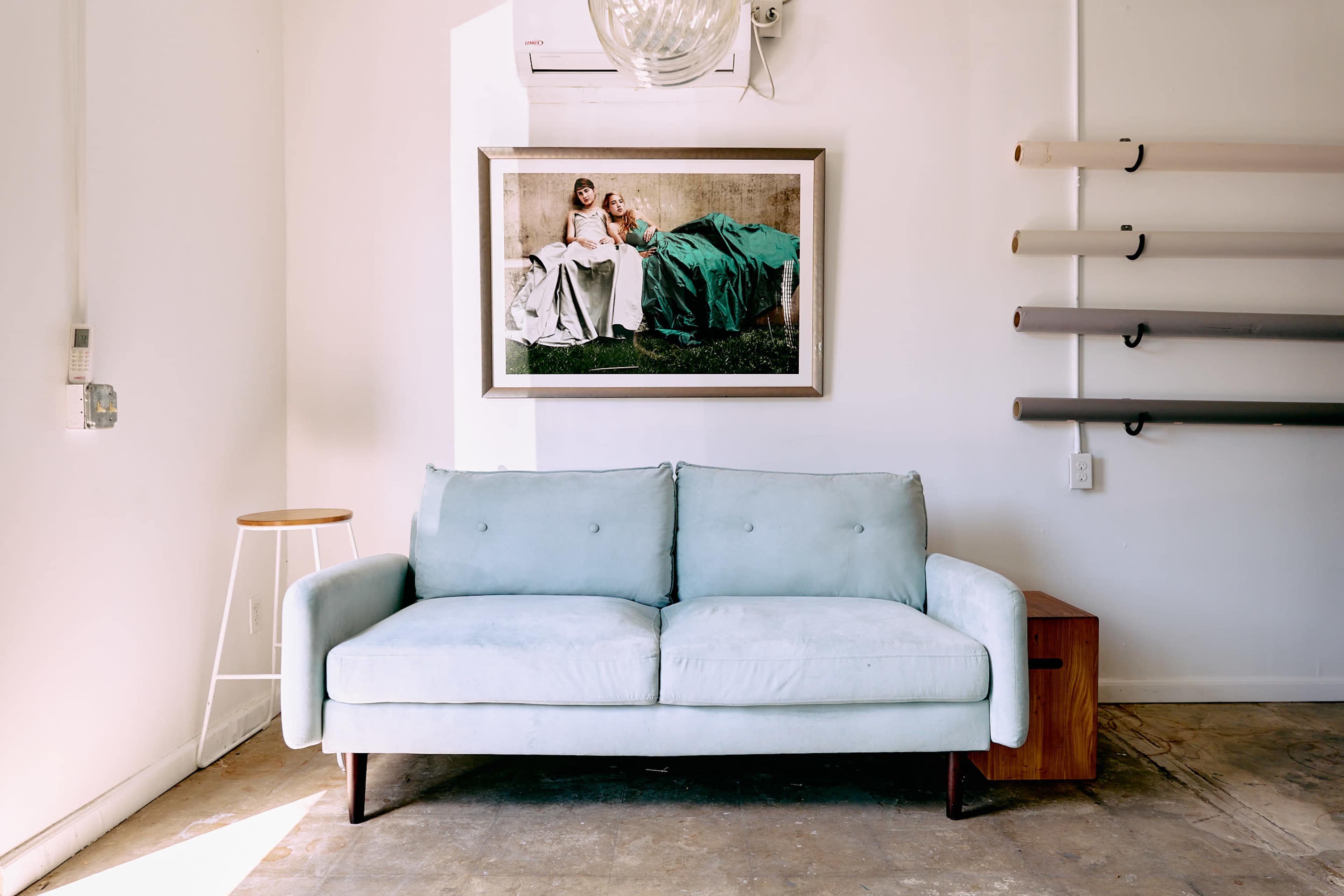 A light blue sofa is positioned in front of a framed artwork on the wall, with a small wooden table and rolled materials displayed to the side.