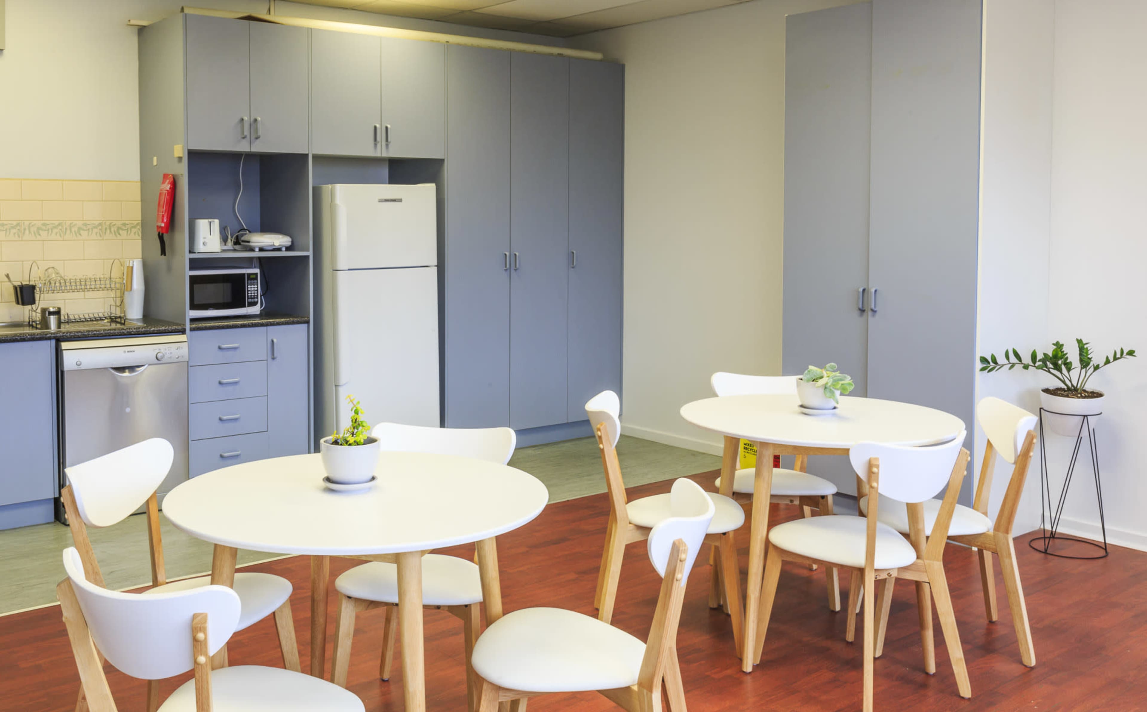 The image depicts a break room with two round tables surrounded by white chairs, a refrigerator, and kitchen appliances against light blue cabinetry.