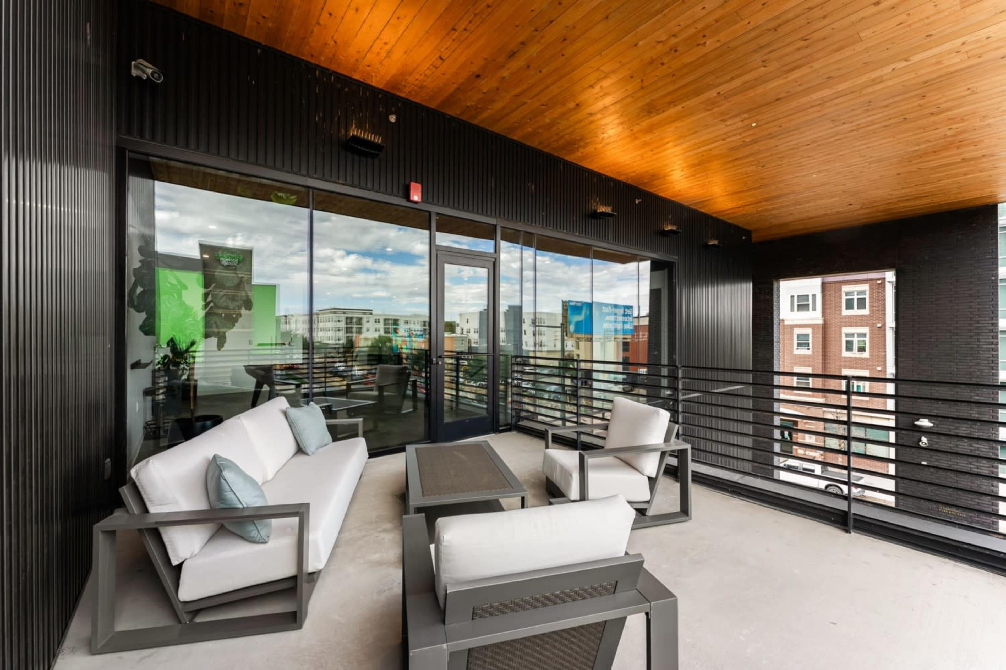 The image shows a modern balcony furnished with white seating and a table, overlooking a cityscape through large glass doors.
