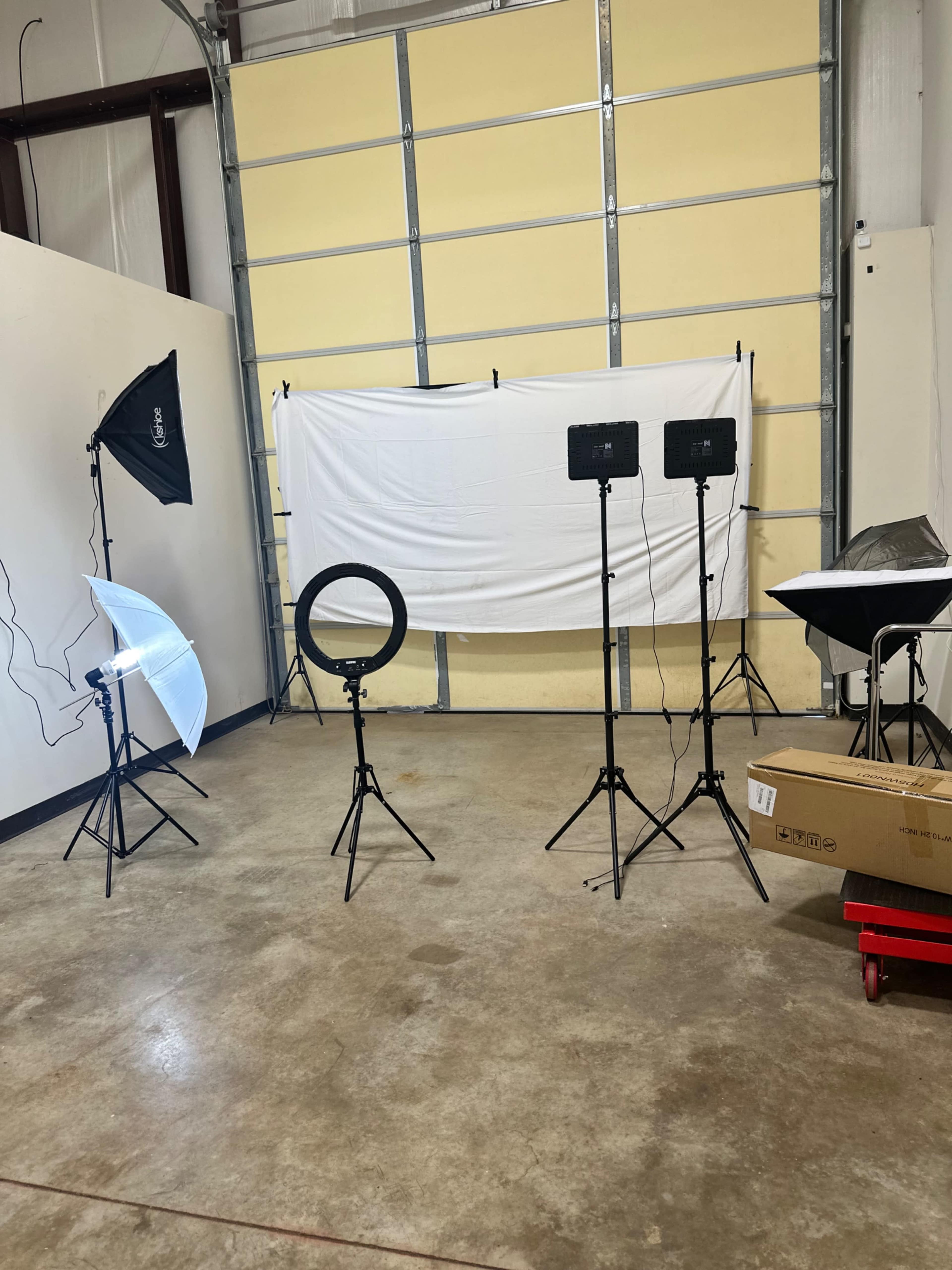 The image shows a photo studio setup with a white backdrop, multiple lighting equipment, and a ring light positioned on tripods.