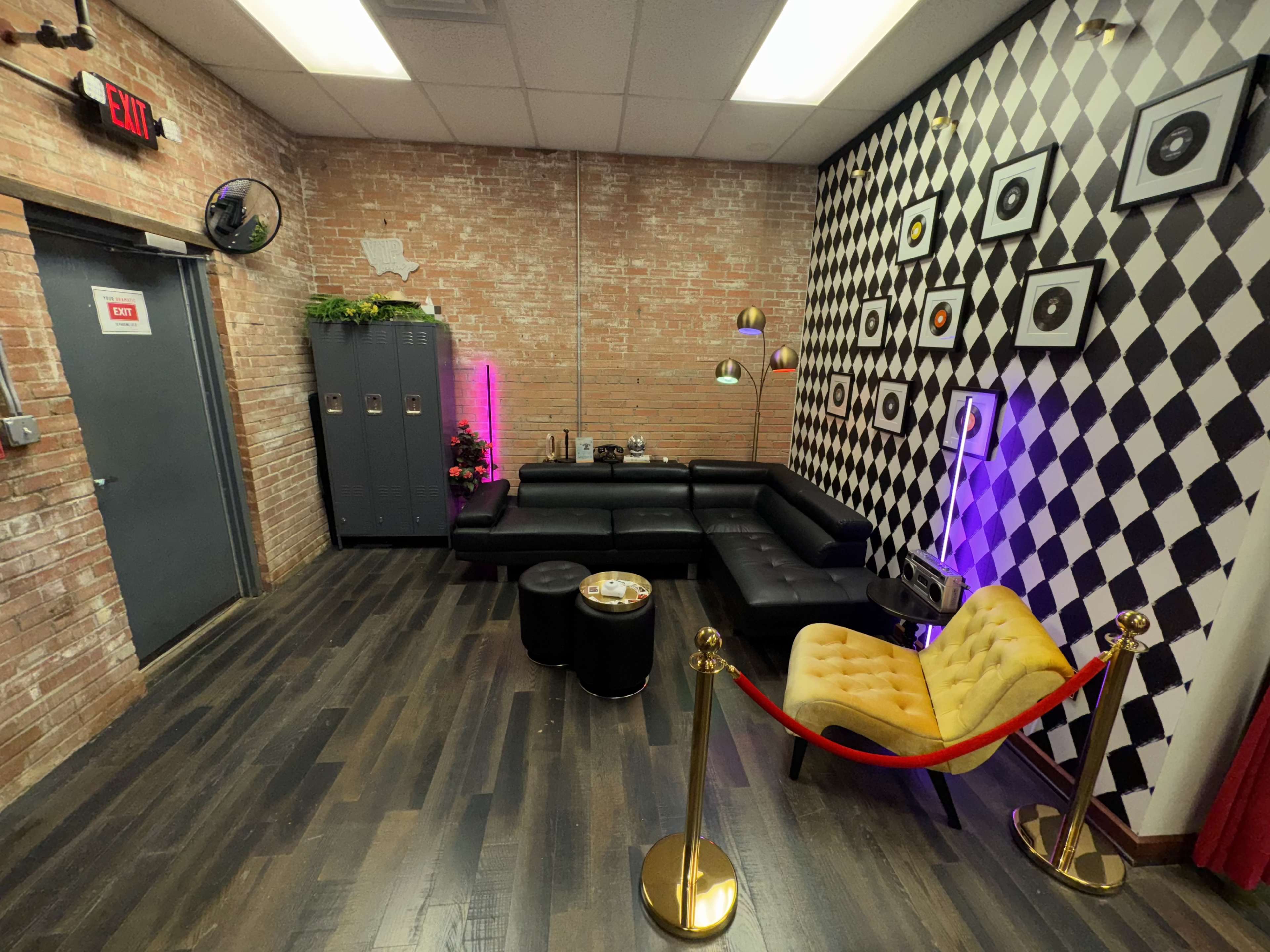 The image shows a modern lounge area with a black and white checkered wall, brick accents, a black sectional sofa, yellow armchair, and decorative lockers.