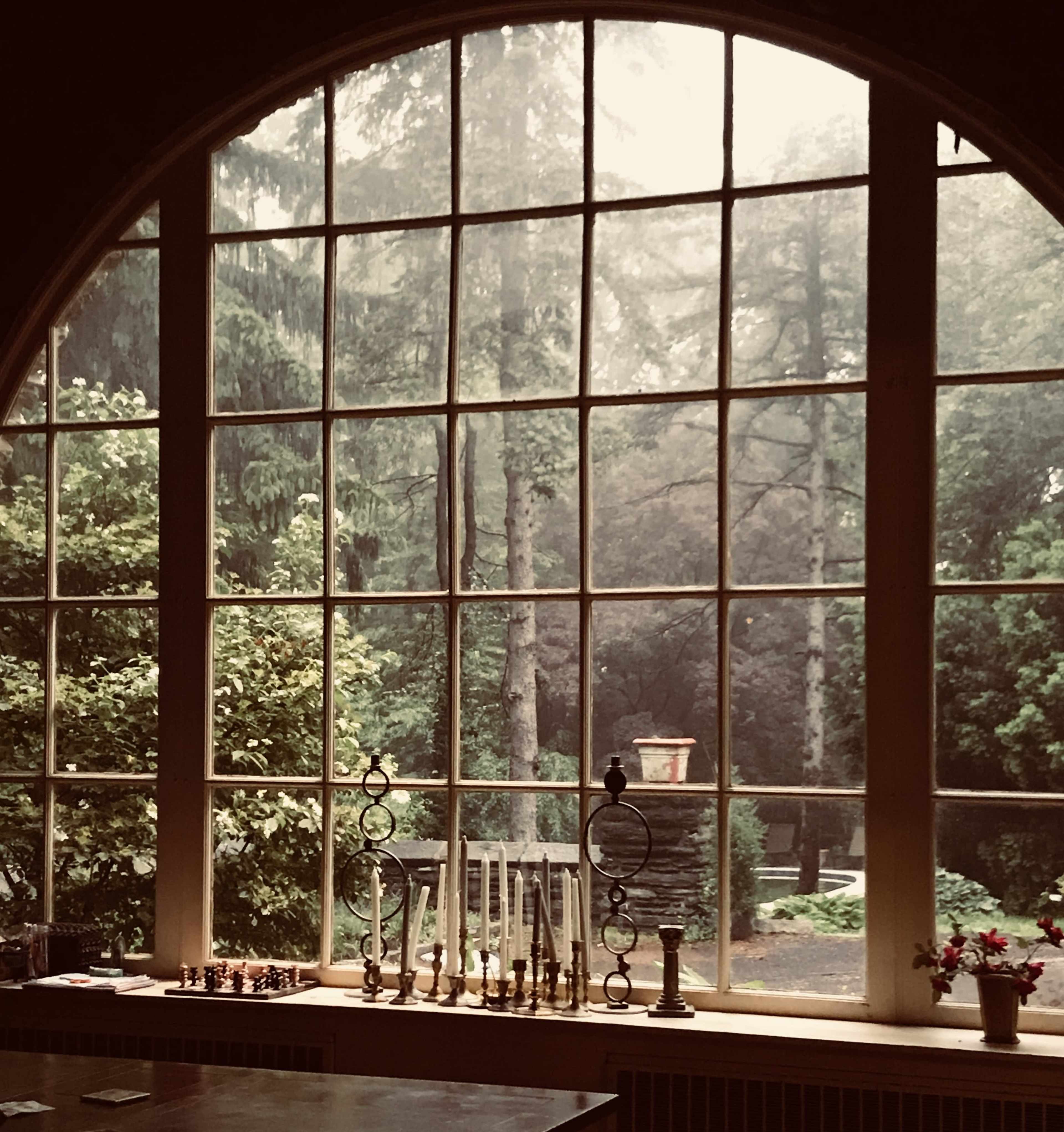 A large arched window frames a misty view of trees and a garden, while various objects sit on a table in the foreground.