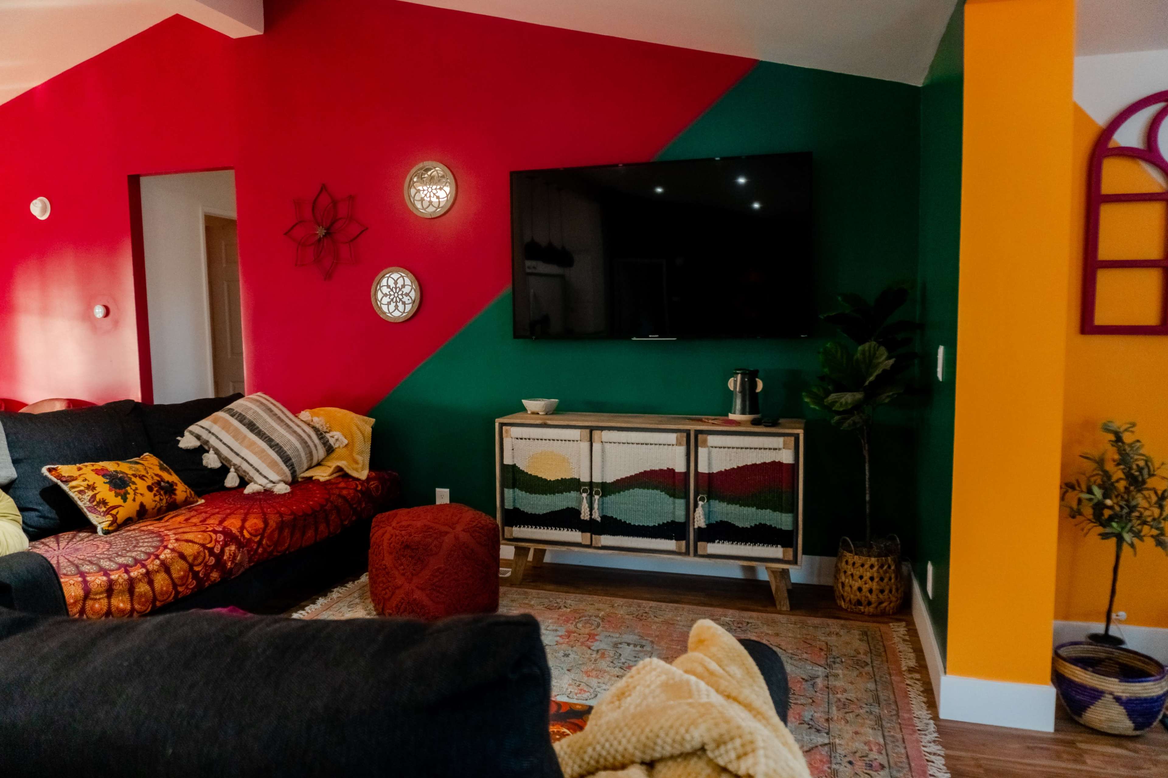 A living room features a dark sofa, colorful accent walls, a television mounted above a wooden cabinet, and decorative items like a rug and plants.